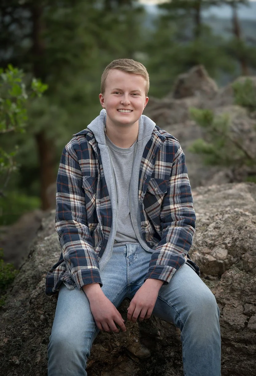 Natural senior portraits while sitting on rocks outdoors, wearing a plaid jacket over a gray hoodie and light blue jeans, with green trees in the background.