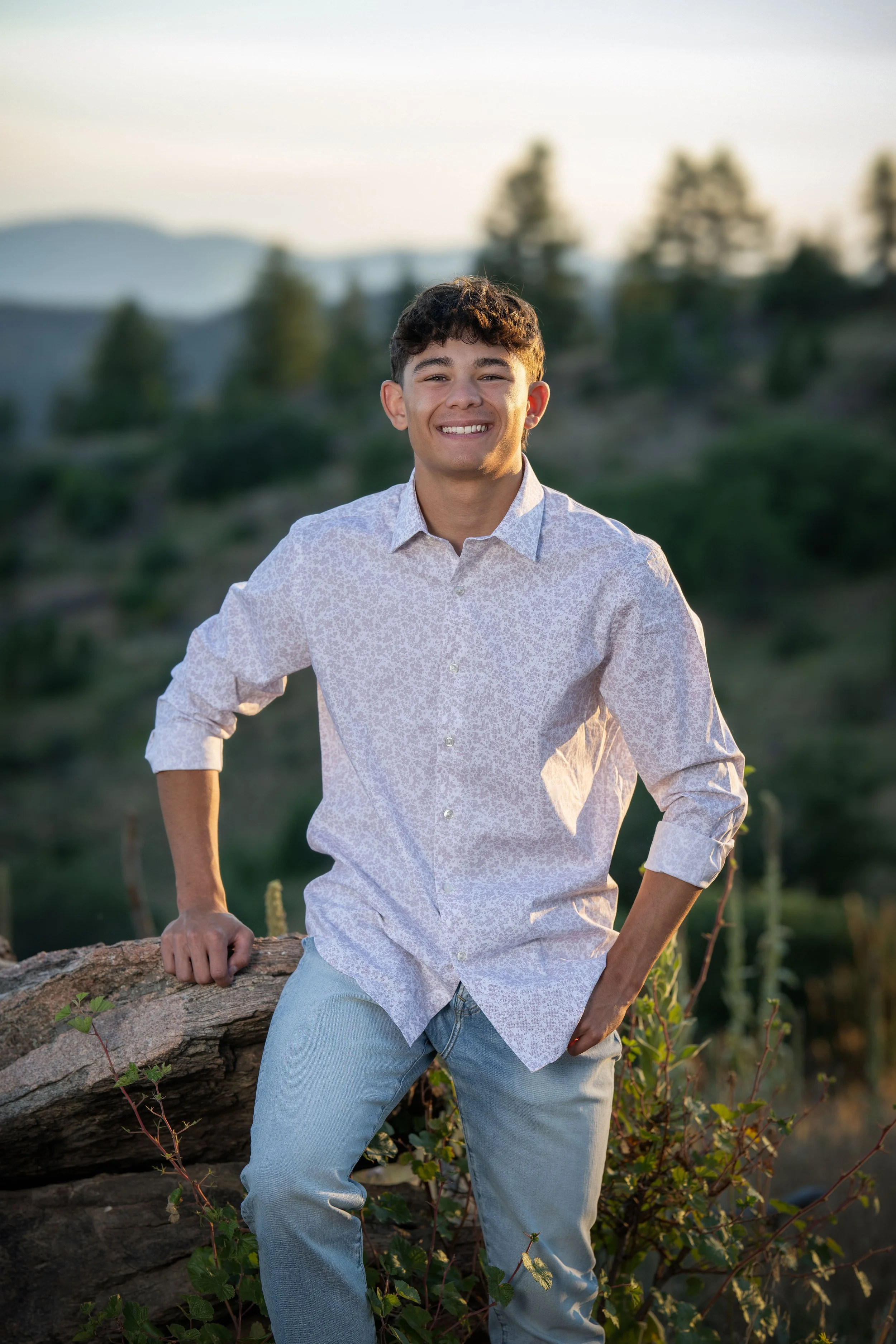 A young man smiling outdoors during sunset, wearing a white patterned shirt and light blue jeans, standing next to a rock and green bushes with a blurred landscape in the background.