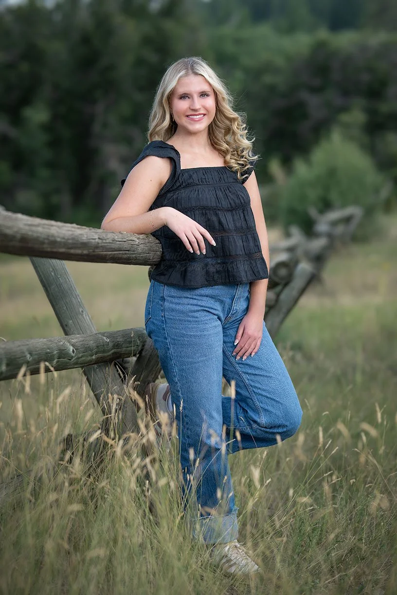 A young woman with long blonde curly hair smiling and leaning against a wooden fence in a grassy outdoor setting.