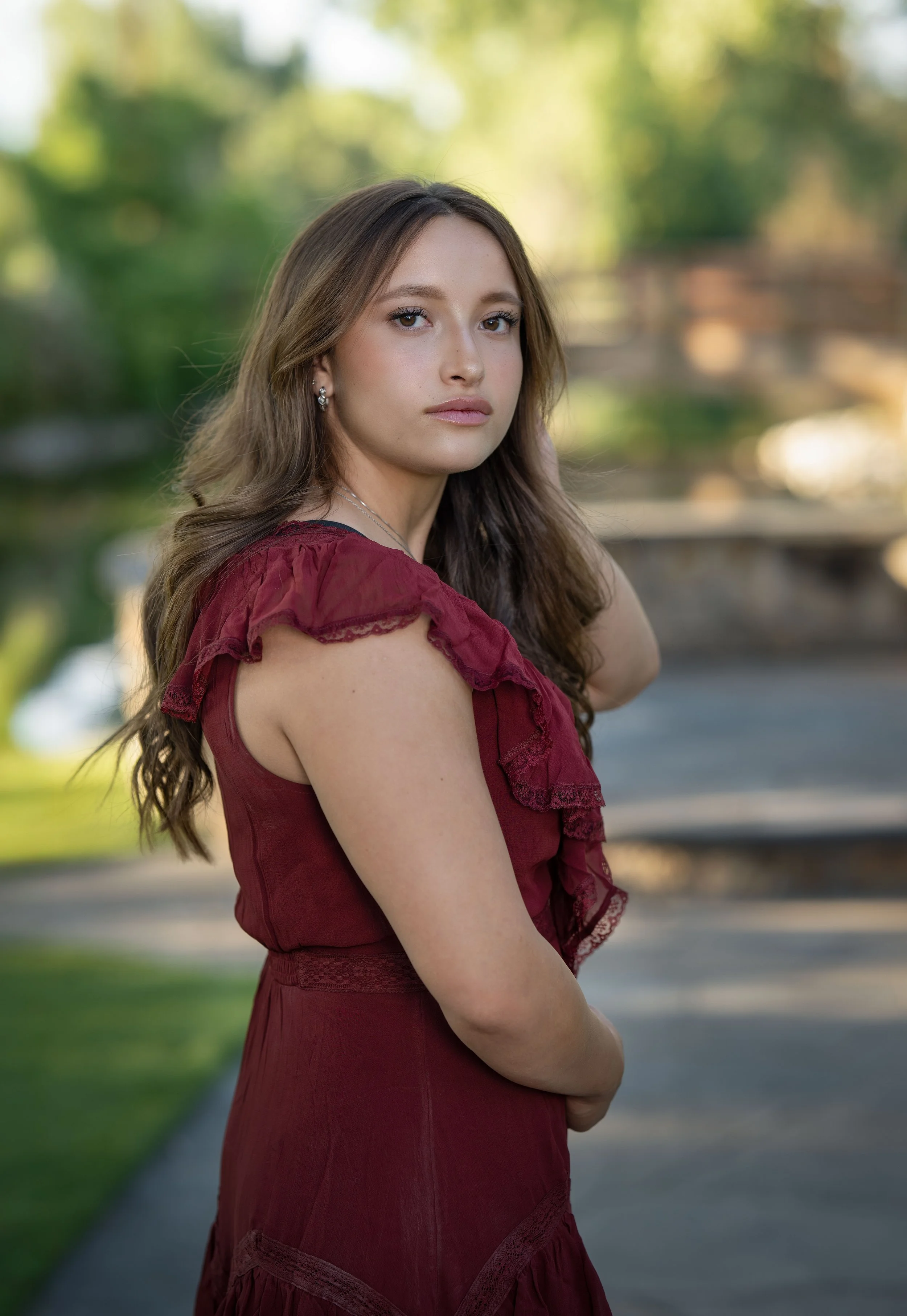 A young woman with long, wavy brown hair and fair skin stands outdoors, wearing a sleeveless dark red dress with ruffled lace details. She has a neutral expression and is looking into the camera, with her left arm bent and hand resting near her waist