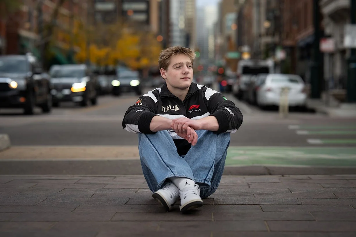 A young man leaning against a brick wall with his left arm raised and resting on a pipe; he is wearing a black jacket, a gray hoodie, a white T-shirt, and beige pants.