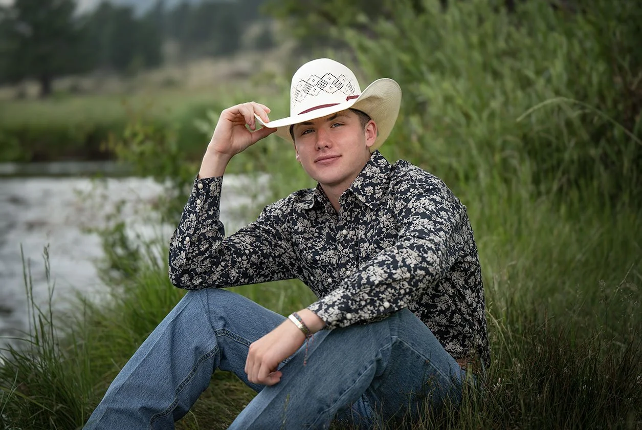 Young man wearing a patterned shirt and jeans sitting outdoors by a river, holding a cowboy hat, with green trees and grass in the background.