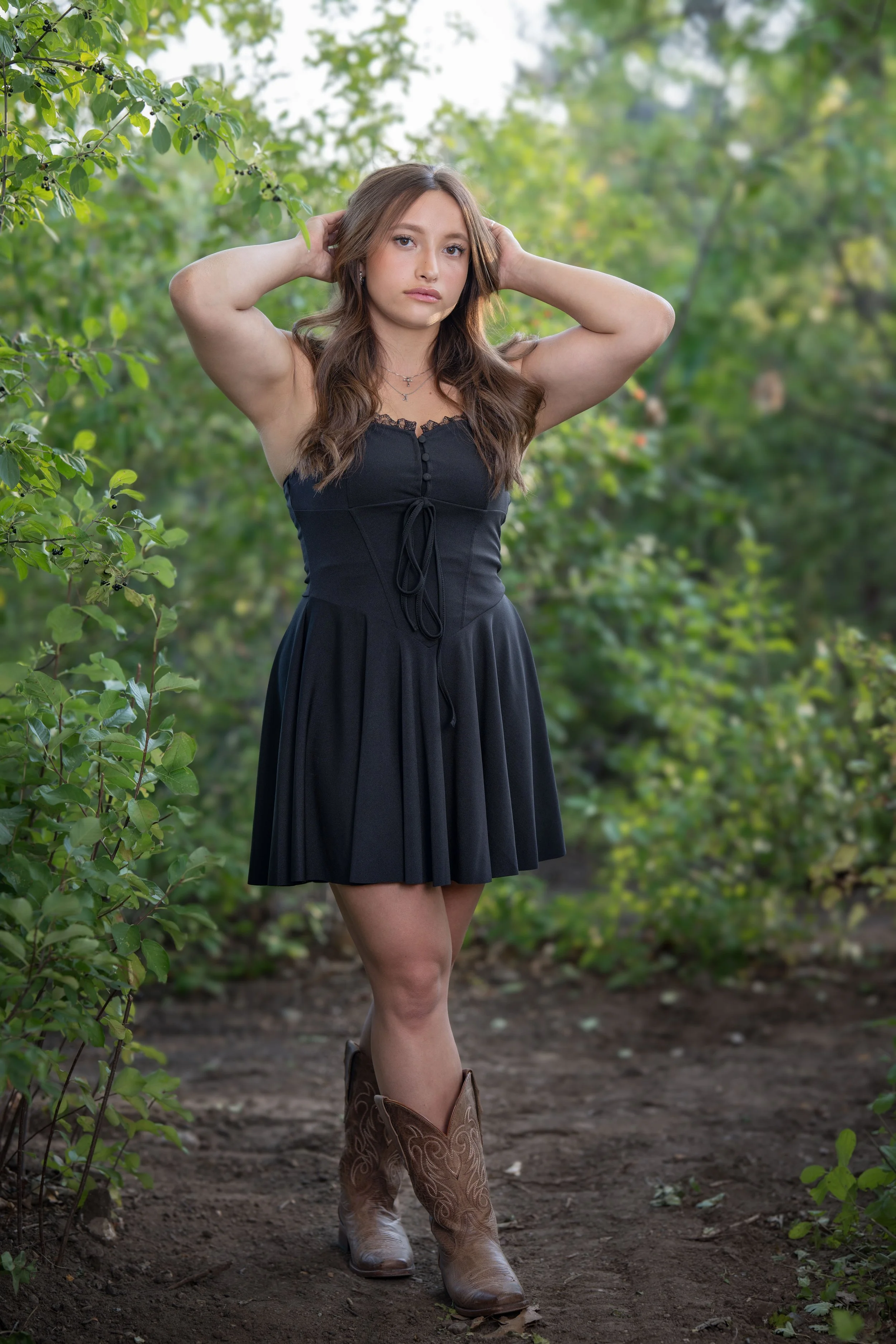 A woman standing outdoors among green bushes, wearing a black dress and cowboy boots, with her hands behind her head and looking at the camera.