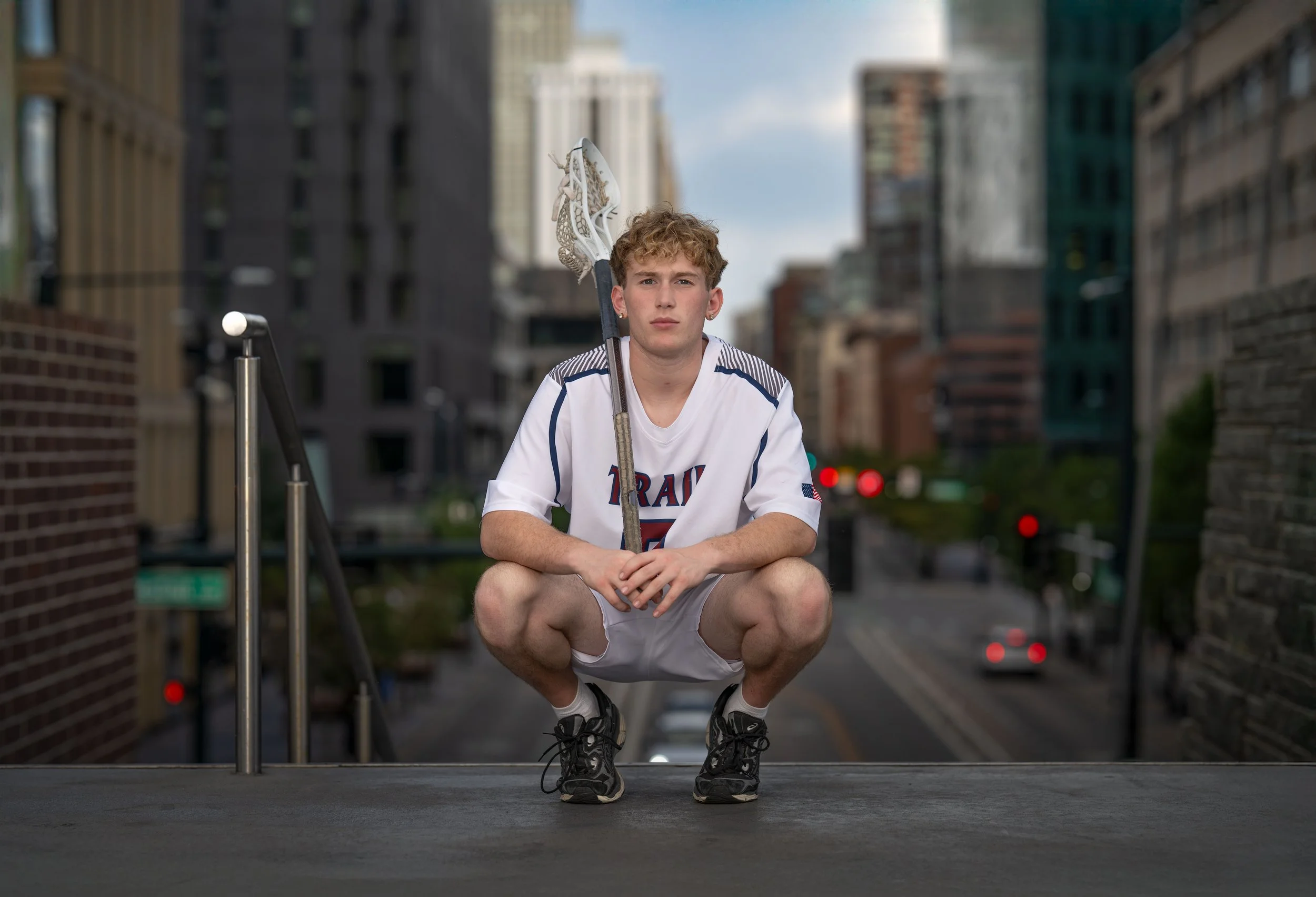 A young male hockey player crouching on a rooftop in an urban setting, holding a hockey stick over his shoulder, wearing a white jersey with blue accents and black athletic shoes.