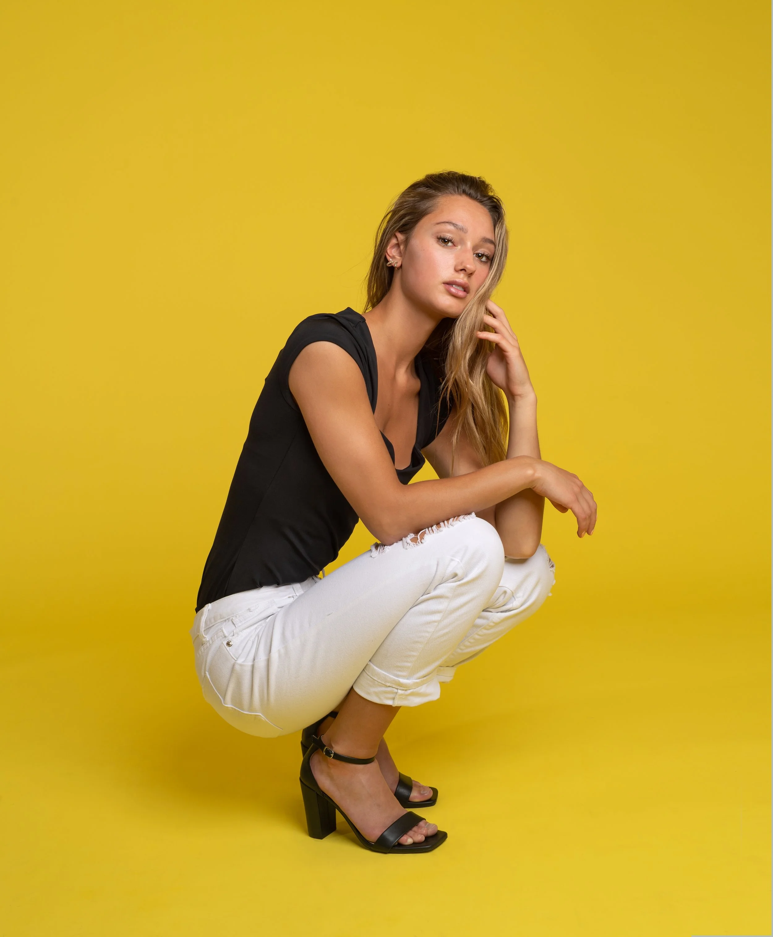 A young woman crouching against a bright yellow background, wearing a black top, white ripped jeans, and black high-heeled sandals.