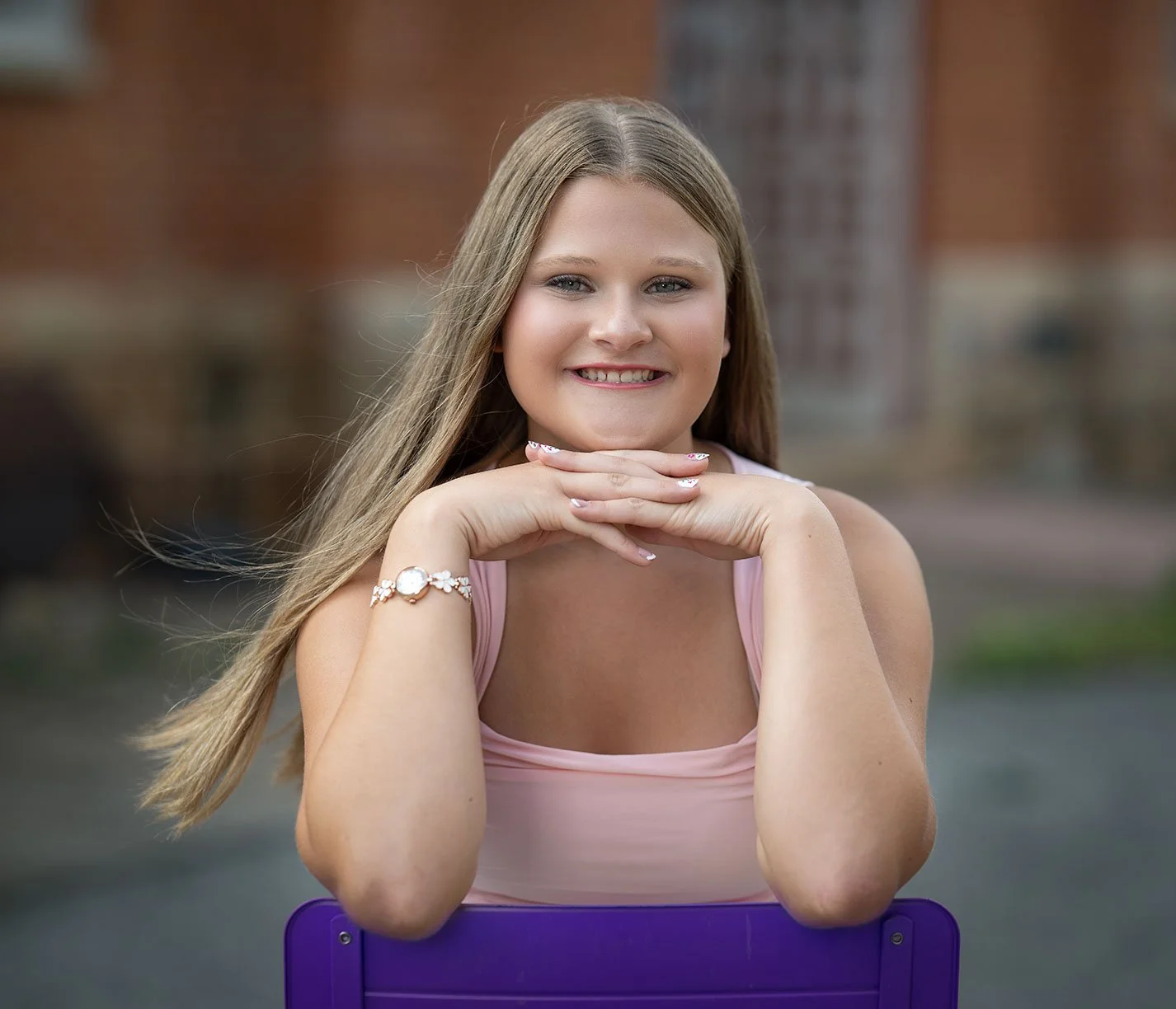 A young woman with long blonde hair smiling, resting her chin on her hands, sitting outdoors with a blurred brick building in the background.