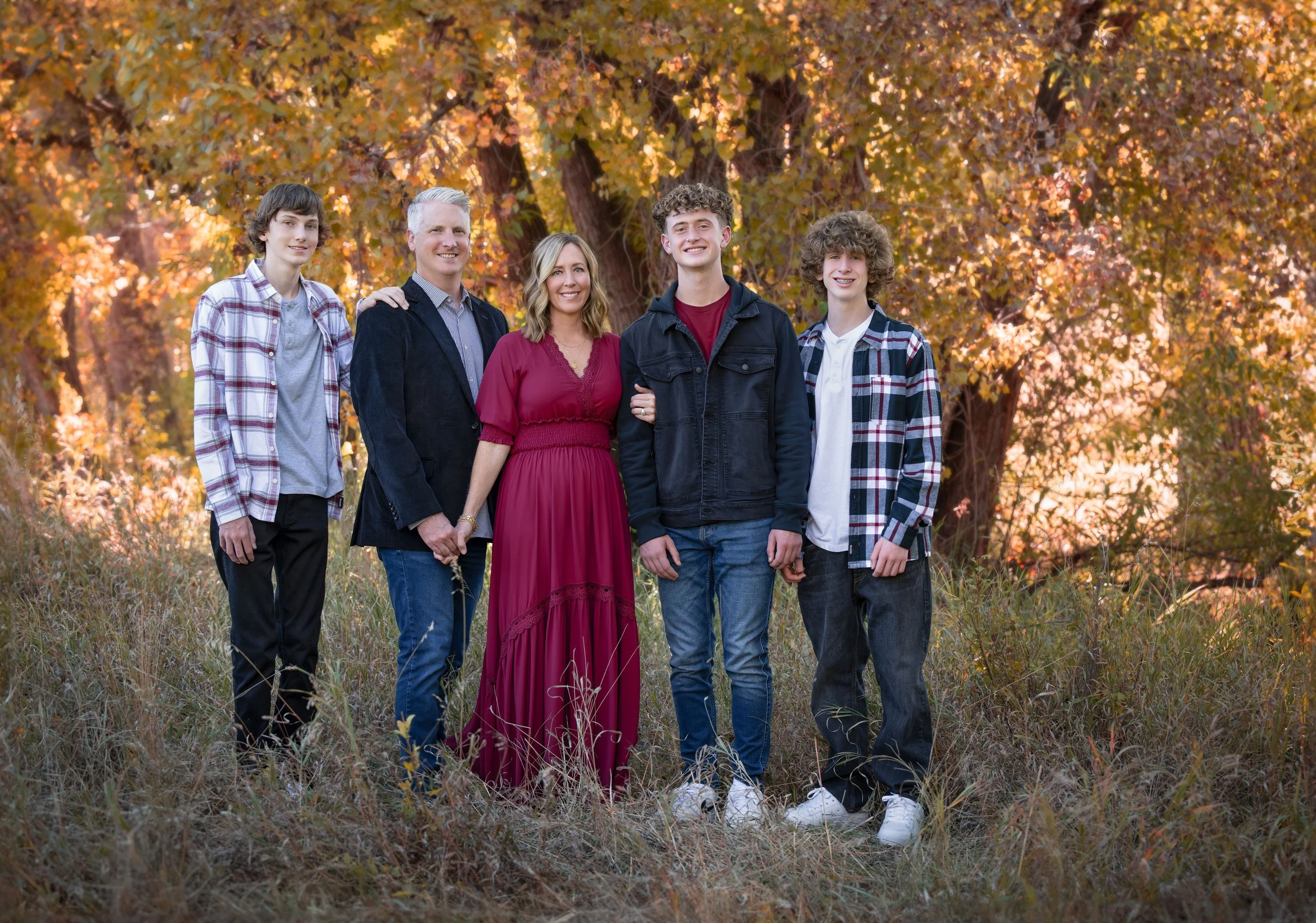 A family of five standing outdoors in a grassy field with trees in the background. The family members are smiling and dressed in casual autumn clothing.