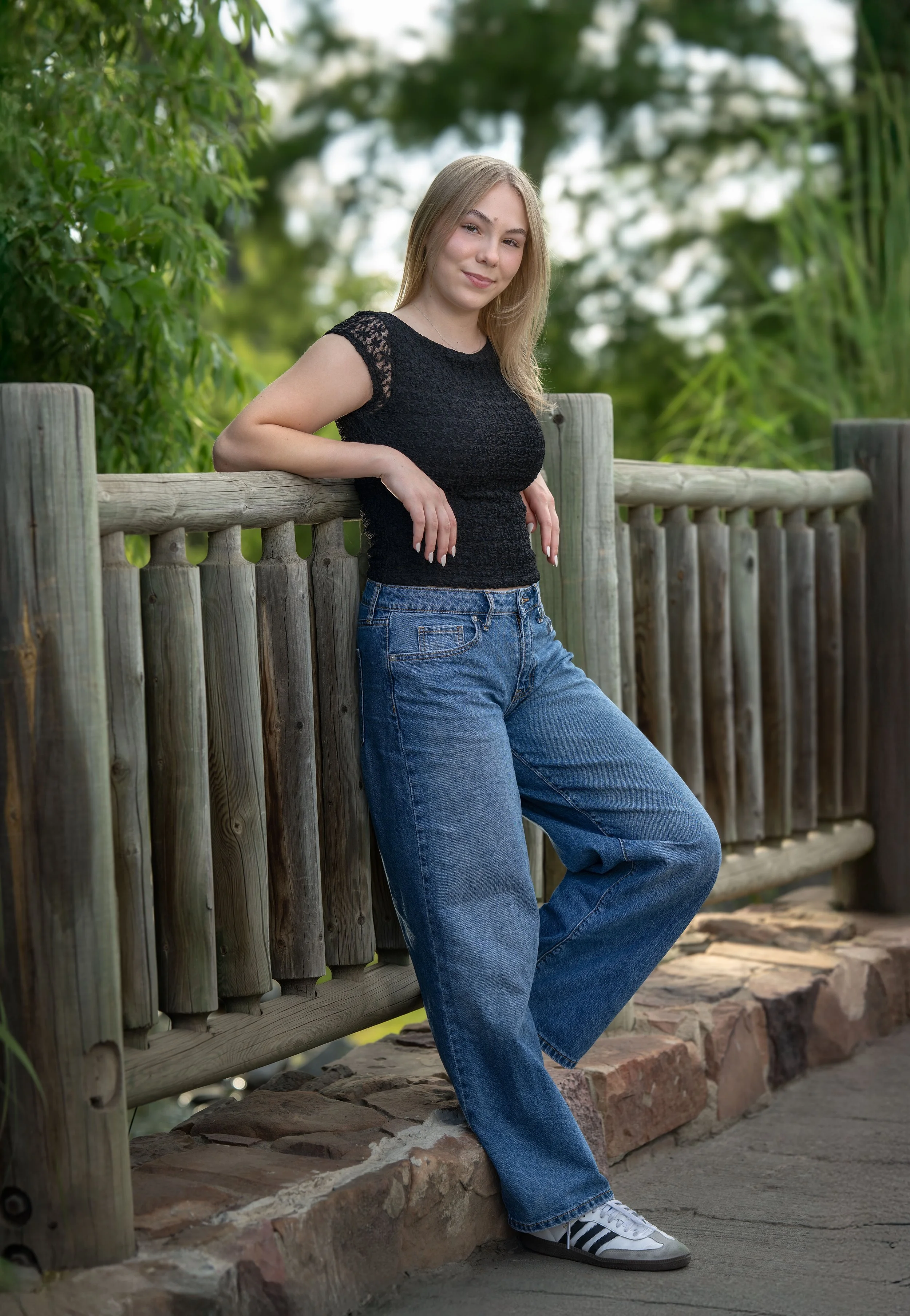 A young woman with blonde hair in casual attire, wearing a black lace top, blue jeans, and white sneakers, leaning against a wooden railing outdoors with green foliage in the background.