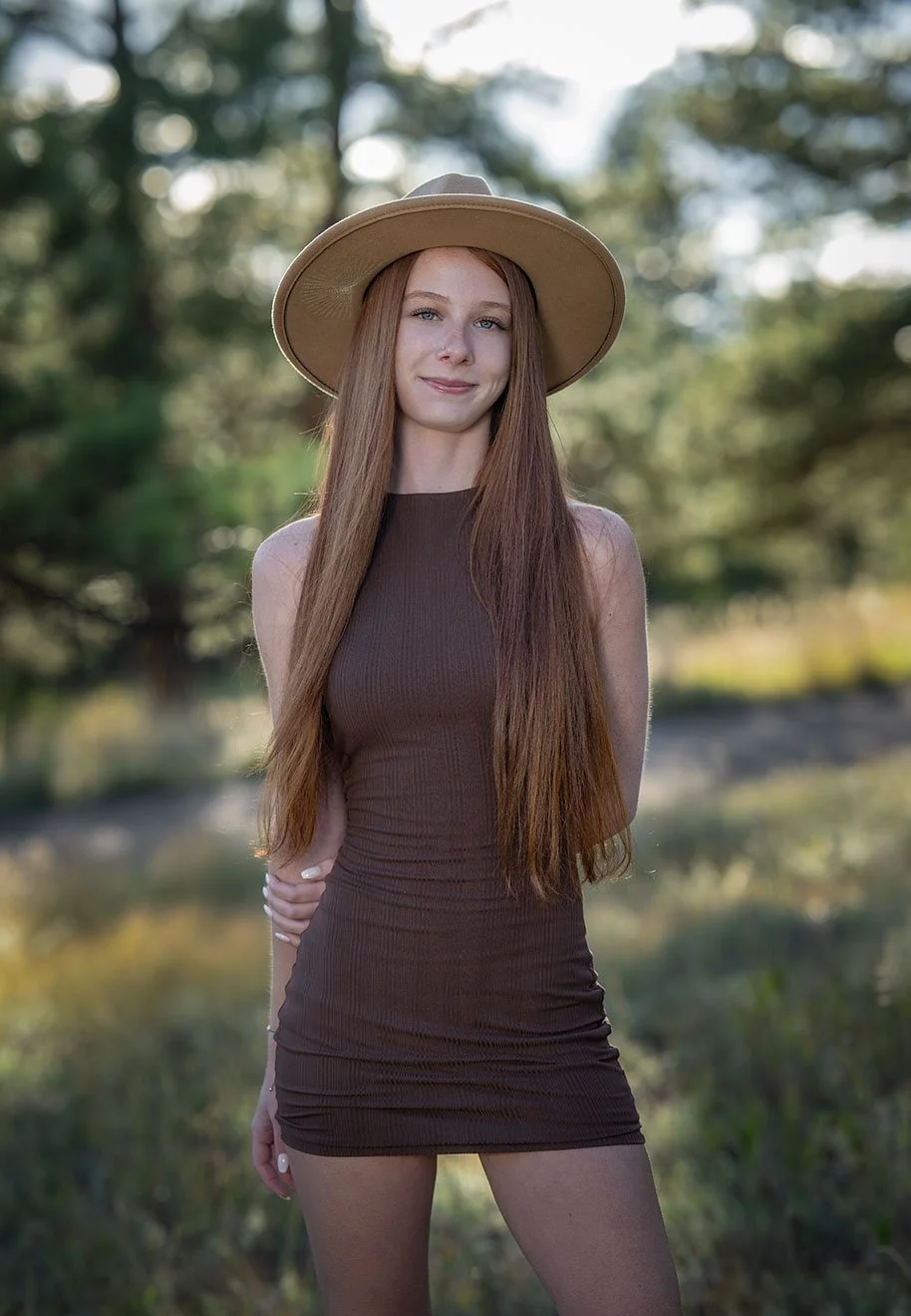 A young woman with long red hair wearing a brown sleeveless dress and a beige wide-brimmed hat, standing outdoors with trees and a blurred natural background.