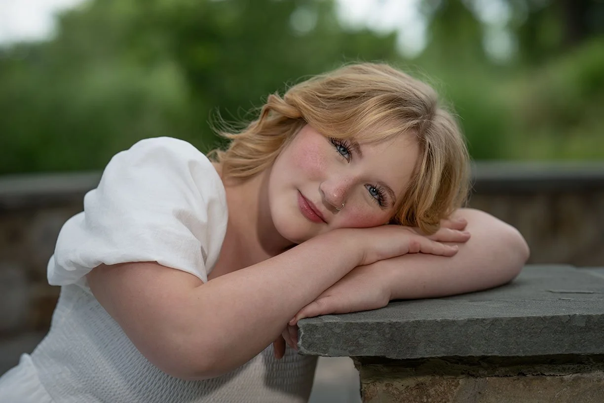 A young woman with short red hair and a septum piercing smiling while resting her head on her crossed arms on a stone surface outdoors.