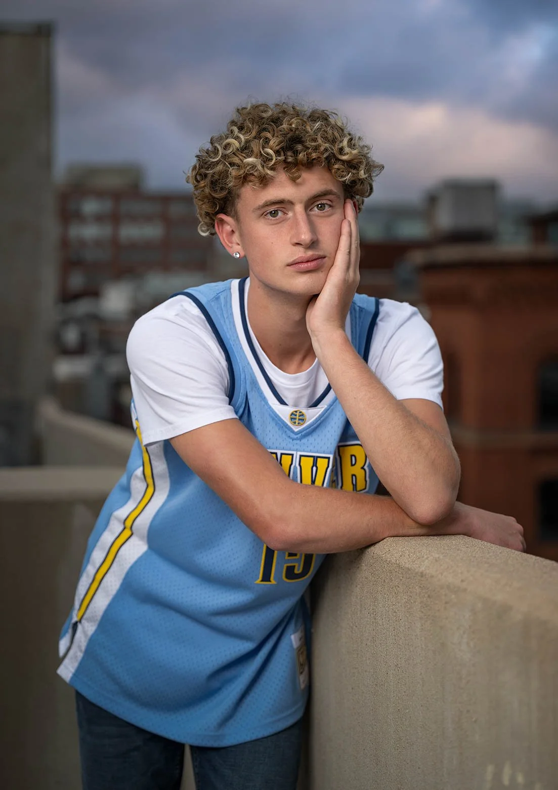 Young person with curly blonde hair wearing a blue sports jersey, leaning on a ledge in an urban rooftop setting during dusk.