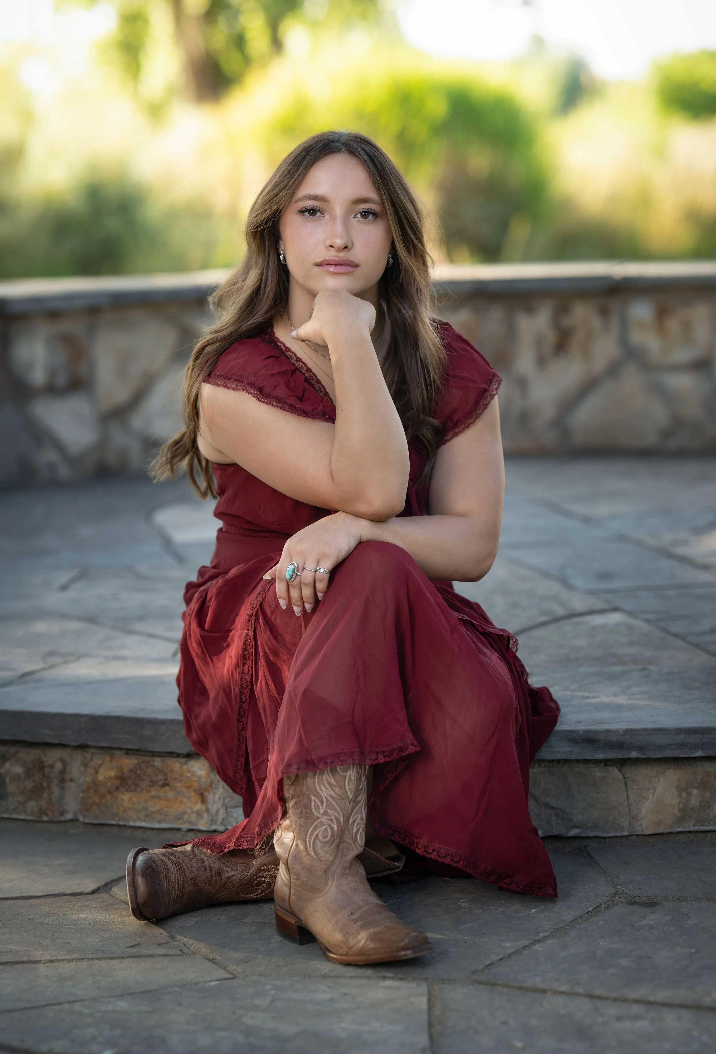 A woman sitting on a stone bench outdoors, wearing a burgundy dress, cowboy boots, and jewelry, with her chin resting on her hand and looking at the camera.