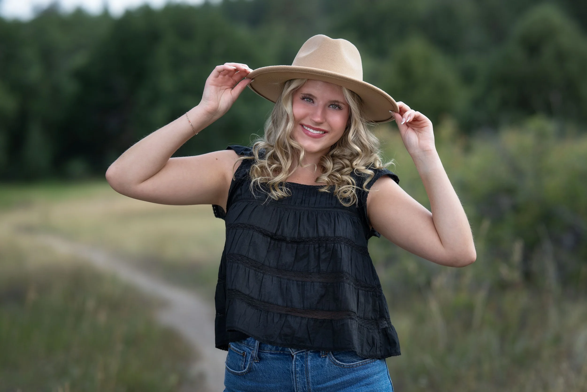 A young woman with curly blonde hair wearing a black sleeveless top and blue jeans, smiling and holding a wide-brimmed tan hat outdoors on a nature trail.