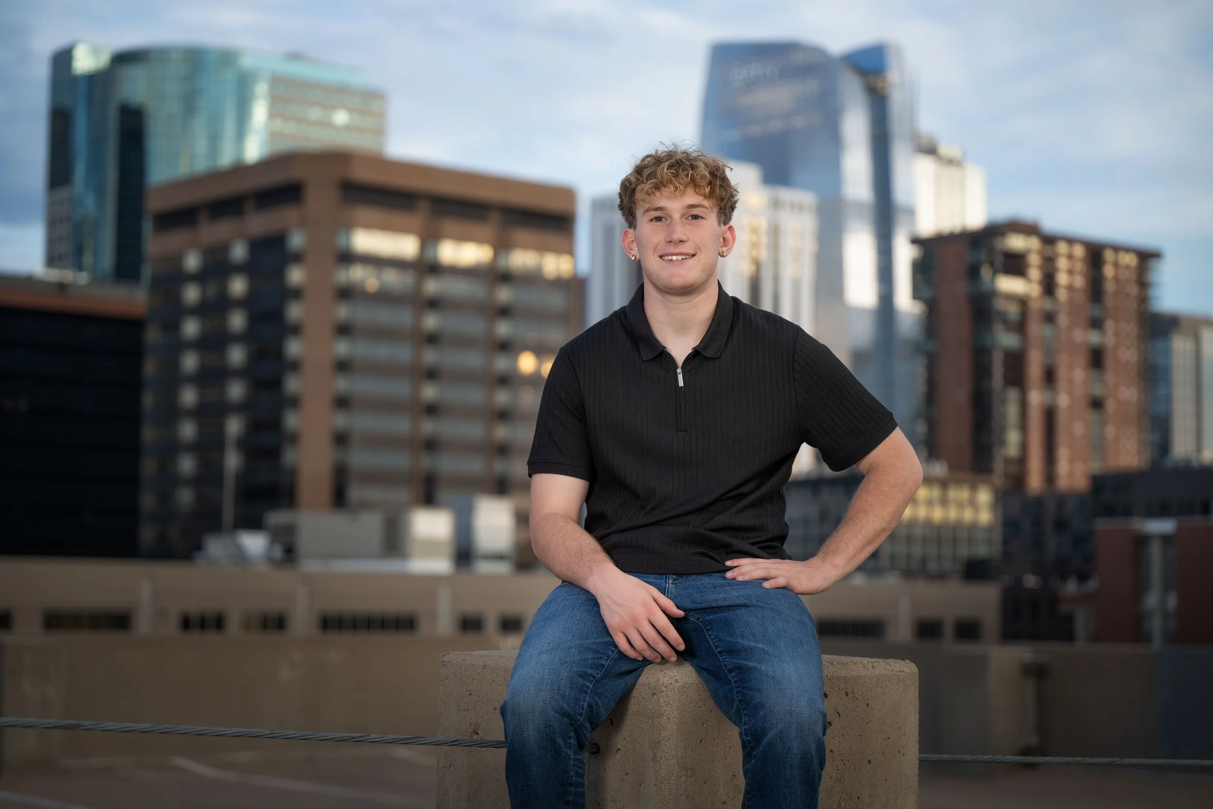A young man with curly hair, wearing a black polo shirt and blue jeans, sitting on a concrete ledge with a city skyline in the background.