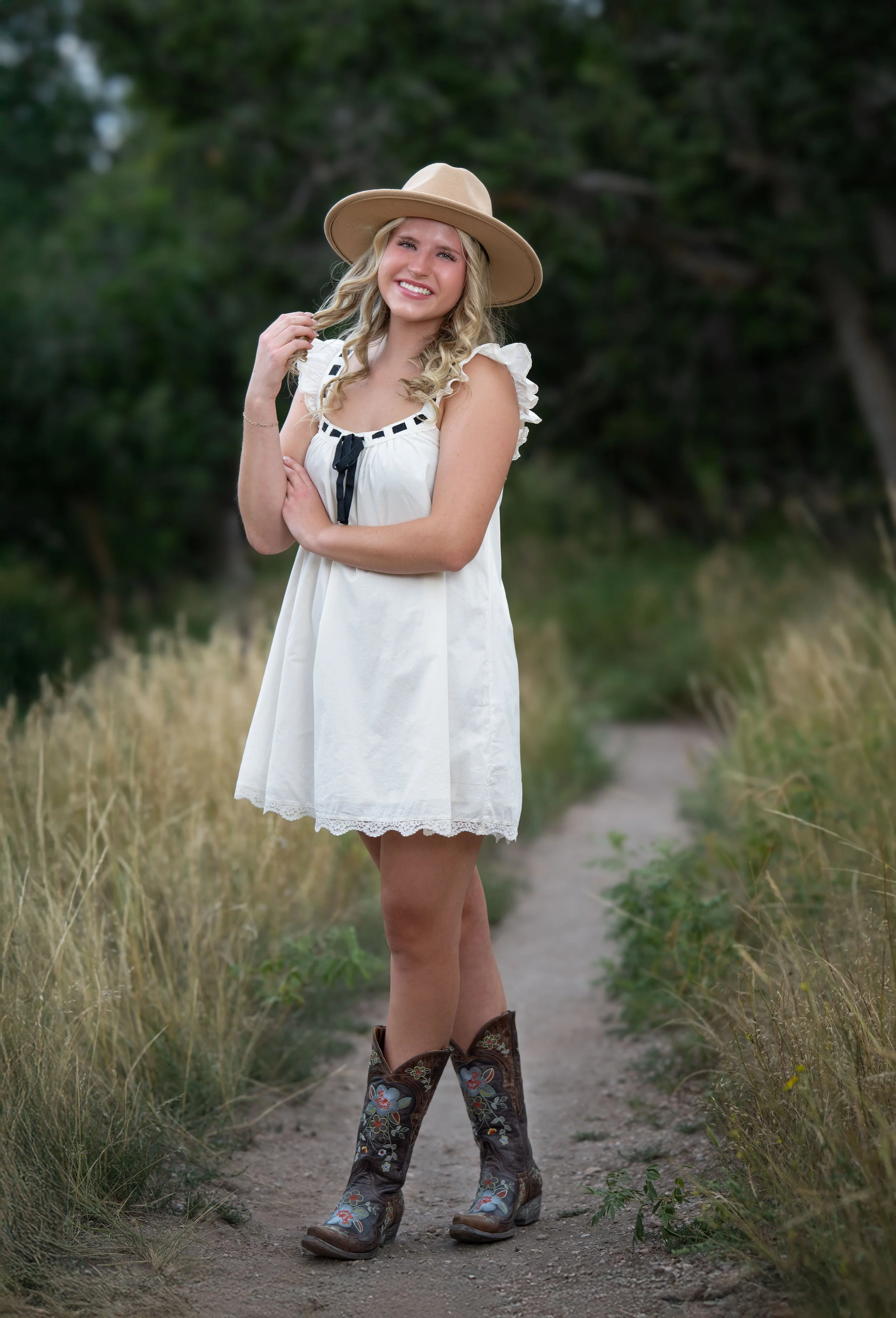 A young woman standing on a dirt path in a green natural setting, smiling, wearing a white dress, cowboy boots with embroidered patterns, and a wide-brimmed beige hat.