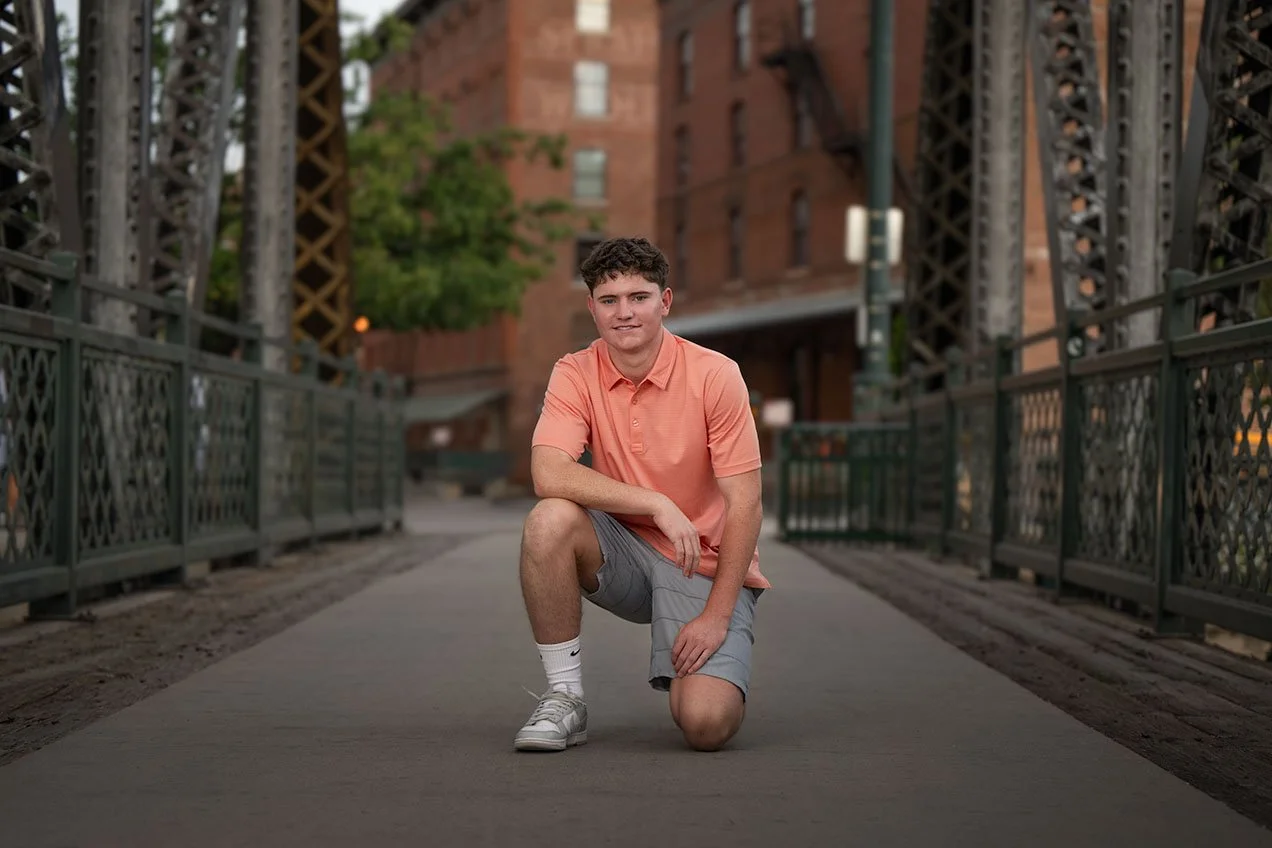 Young man crouching on a bridge with brick buildings and green trees in the background.