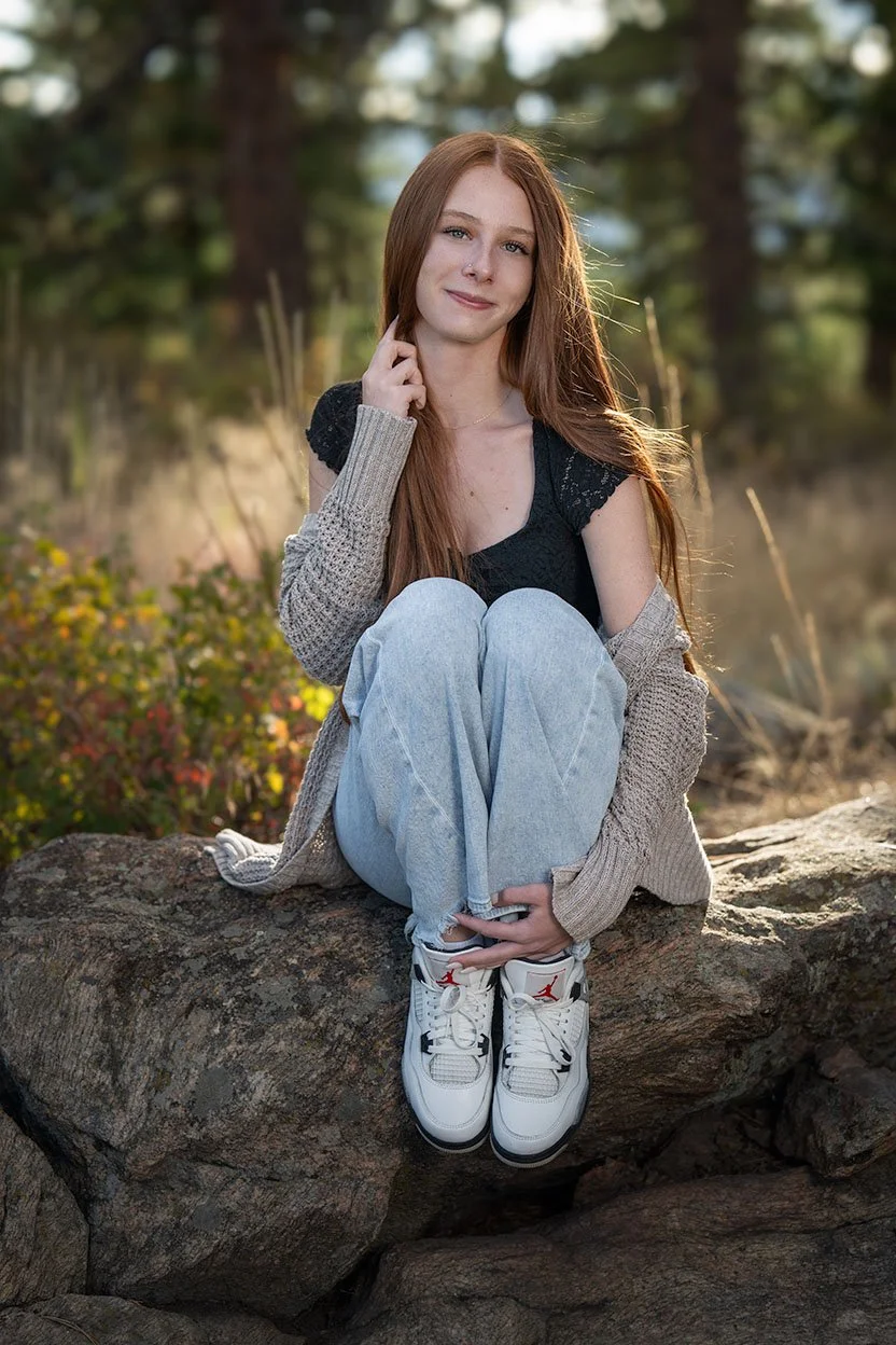 A young woman with long red hair sitting on a rock outdoors, wearing a black top, light blue jeans, white sneakers with red accents, and a gray cardigan, with a forested background.