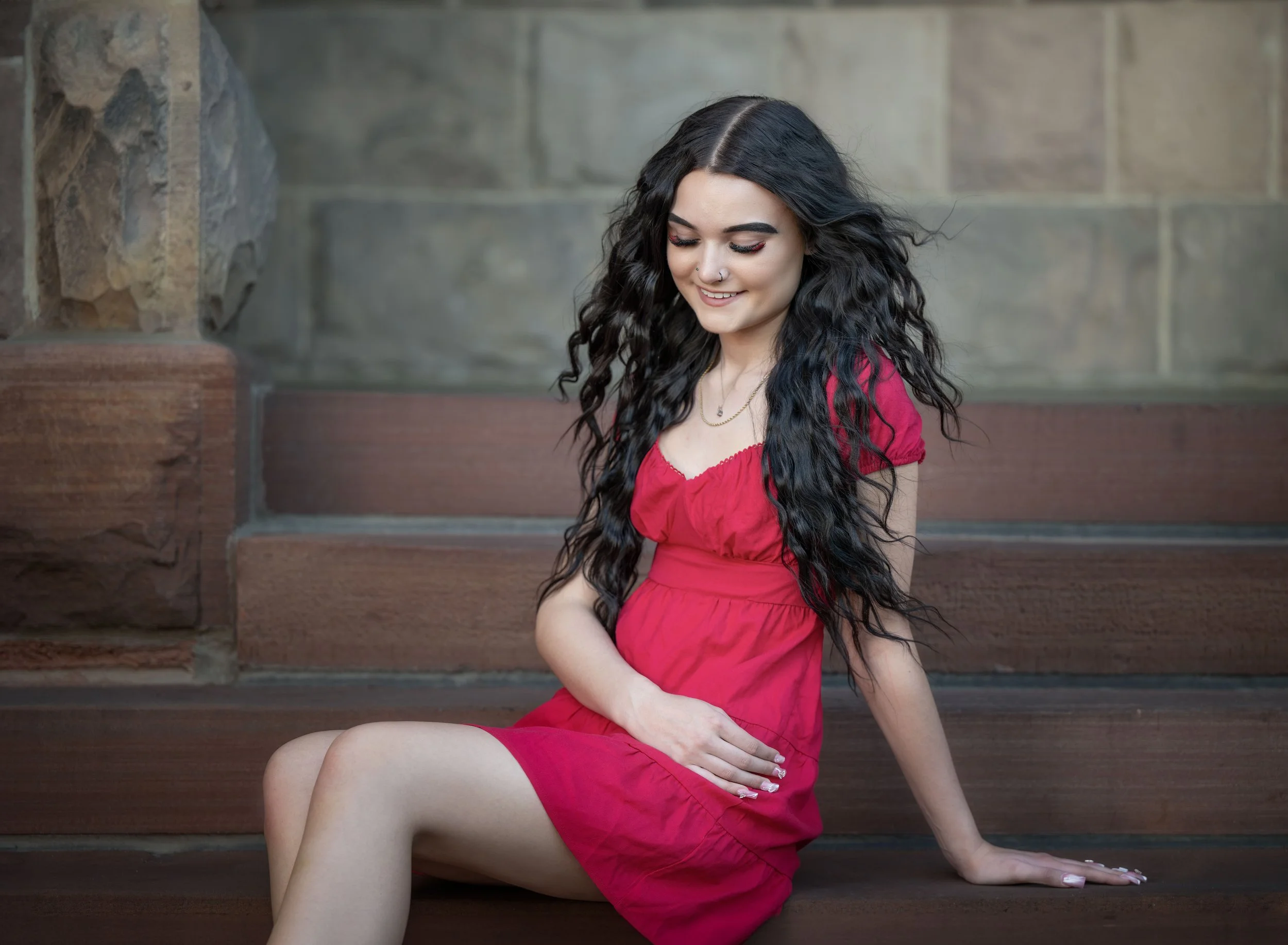 A young woman with long curly black hair, wearing a red dress, sitting on stone steps and smiling.