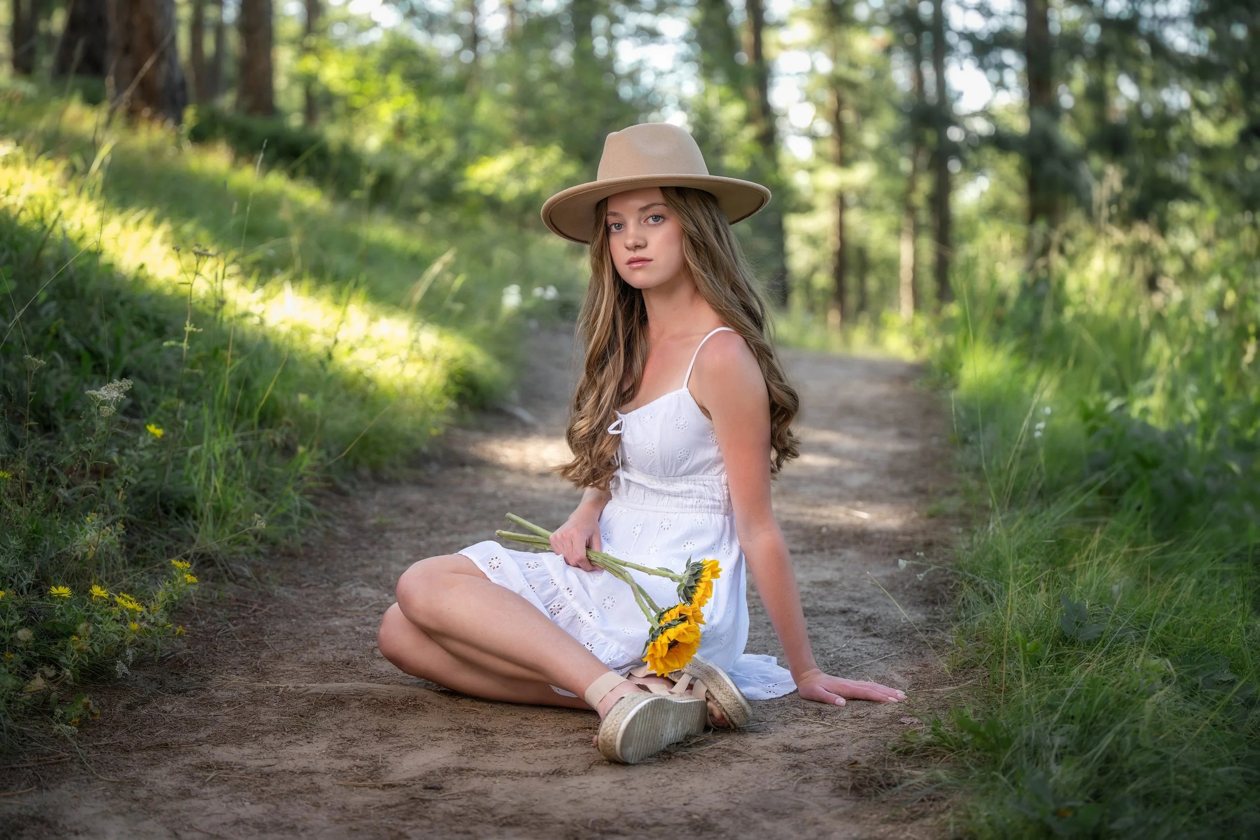 A young woman sitting on a dirt trail in a forest, wearing a white dress and a wide-brimmed hat, holding a bouquet of yellow sunflowers.
