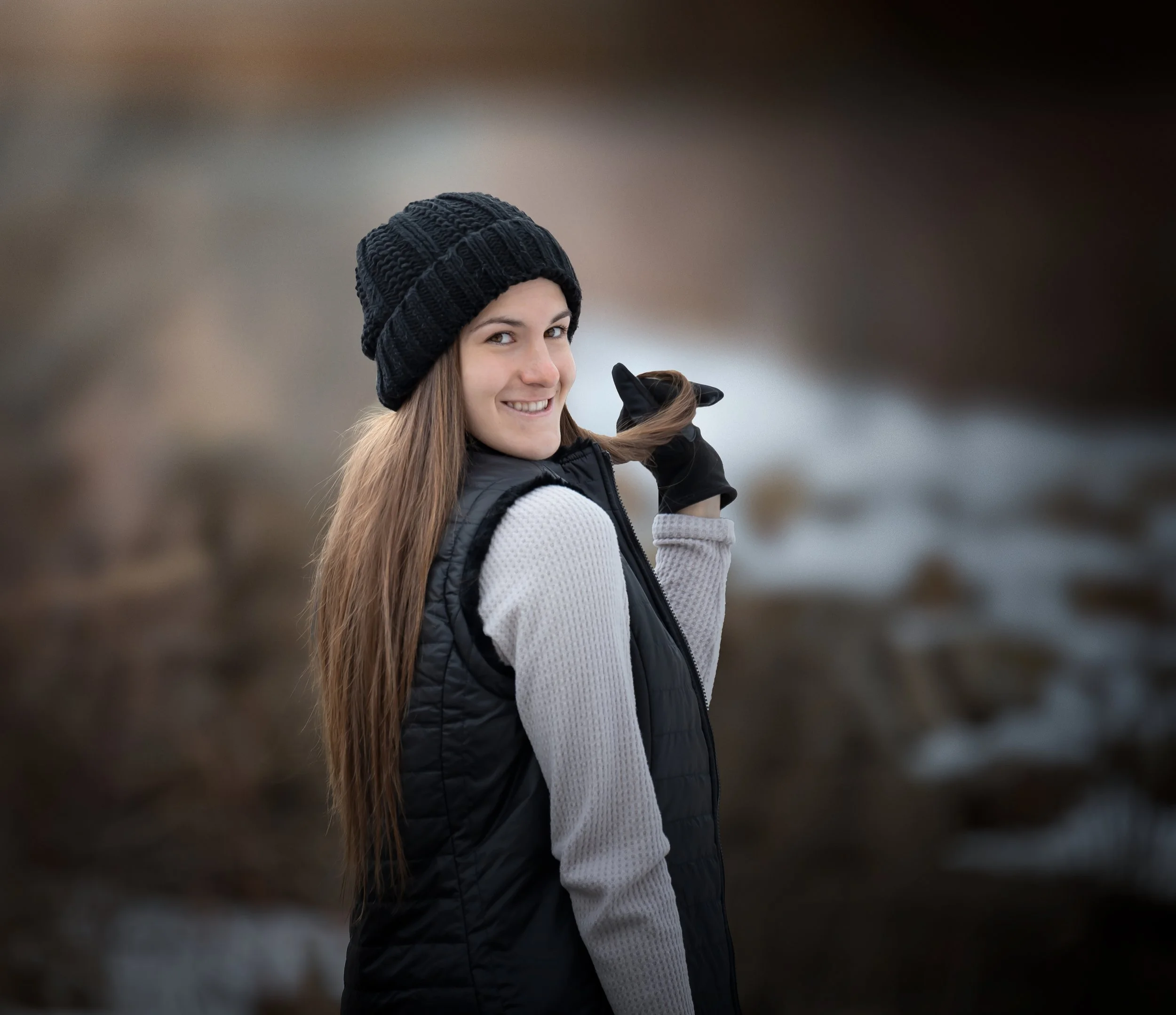 A young woman with long brown hair, wearing a black knit beanie, a gray sweater, a black vest, and black gloves, smiling while standing outdoors near a blurred water body.