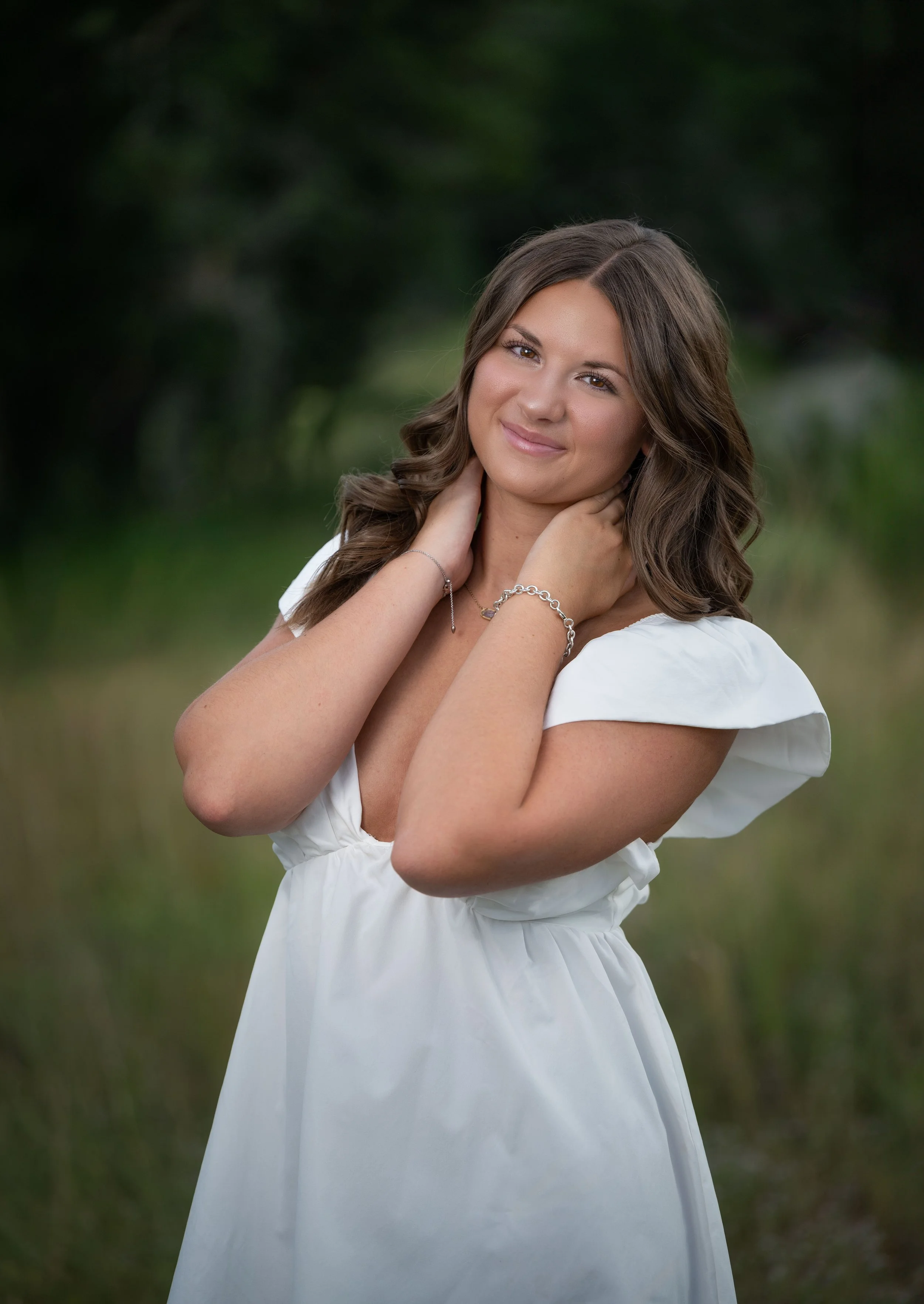 A young woman with long, wavy brown hair, wearing a white dress, standing outdoors with greenery in the background. She is smiling softly, with her hands gently touching her neck.