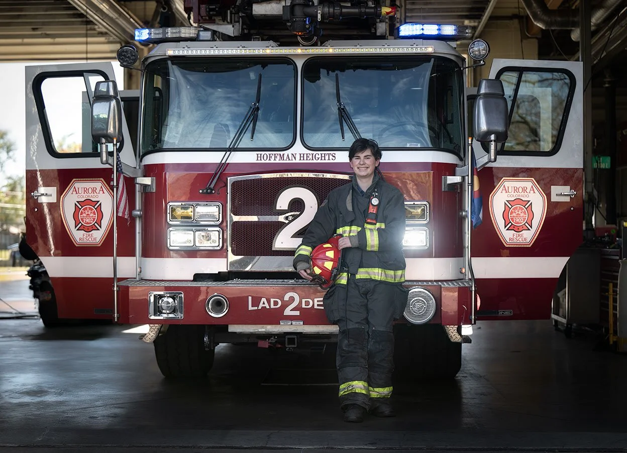 A young firefighter in uniform smiling while standing in front of a red fire truck at a fire station. The fire truck has Aurora Fire Rescue logos and is labeled with the number 2.