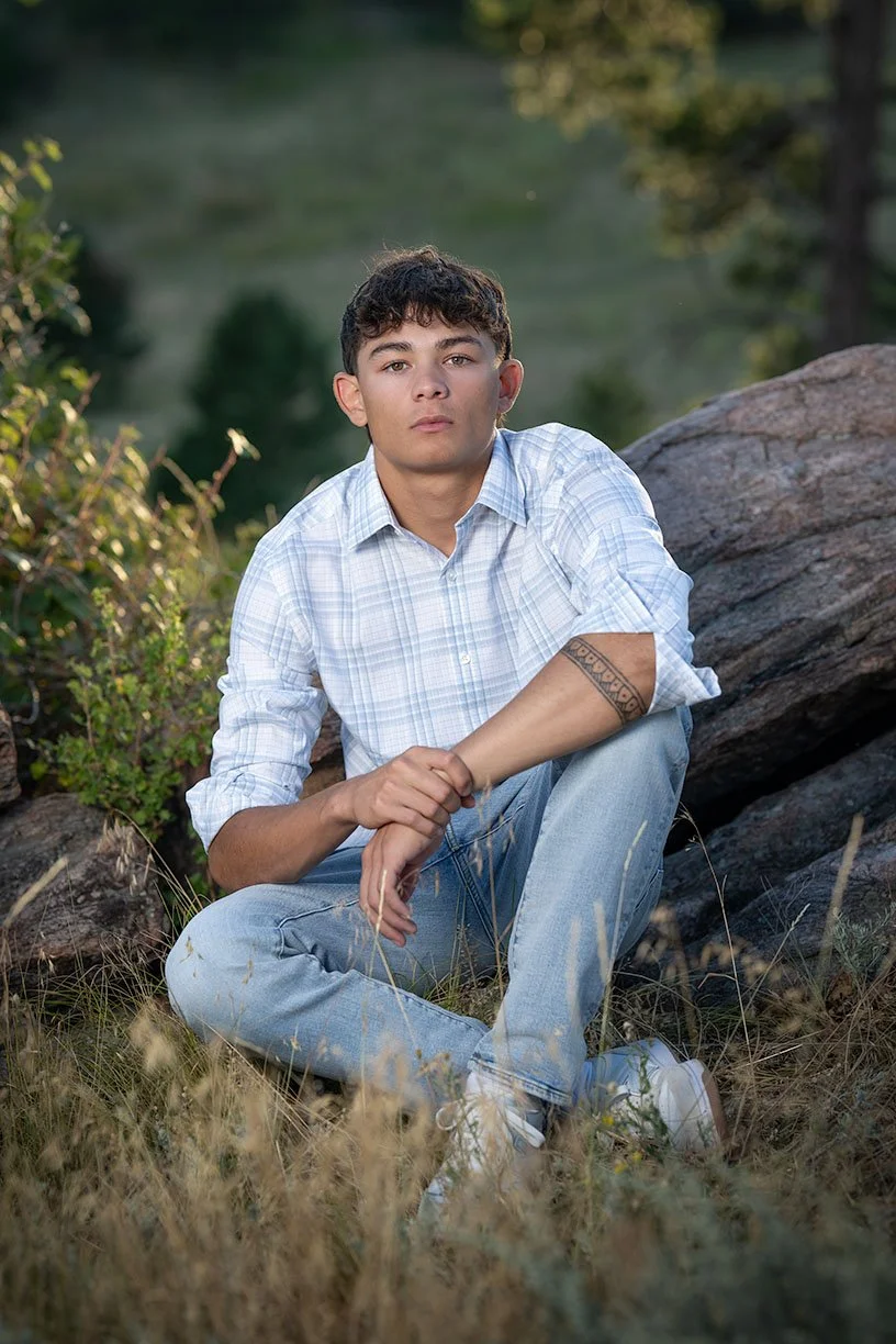 Young man with curly hair sitting on grass near rocks and trees in a natural outdoor setting.