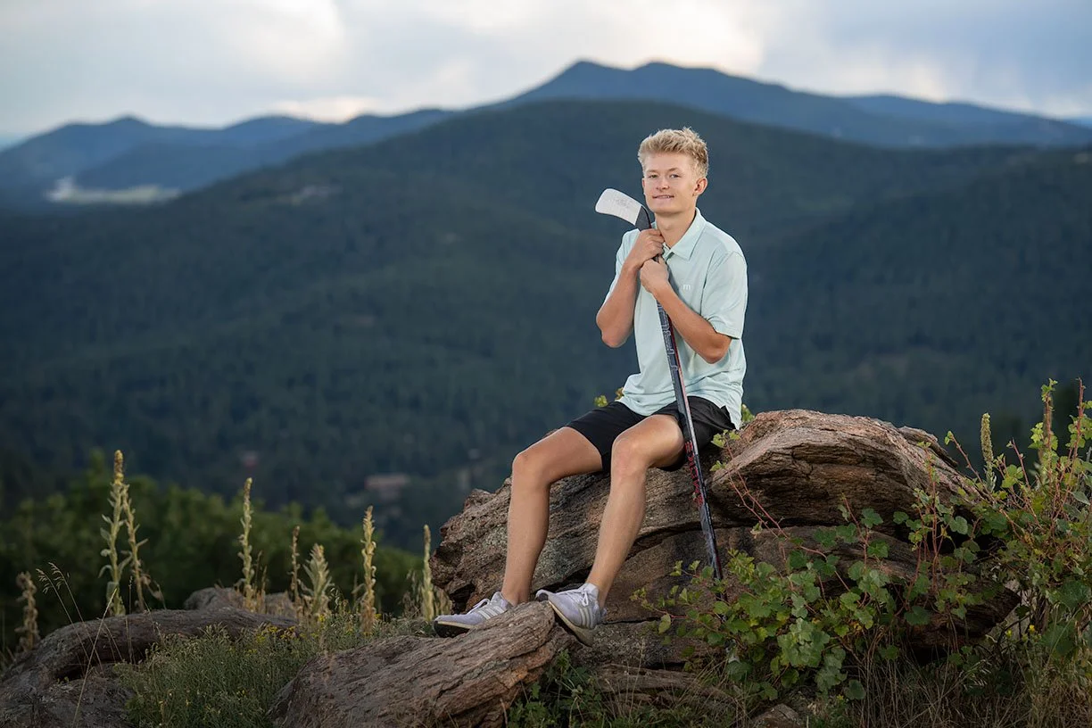A teenage boy sitting on a large rock outdoors, holding a golf club, with a mountain landscape in the background.
