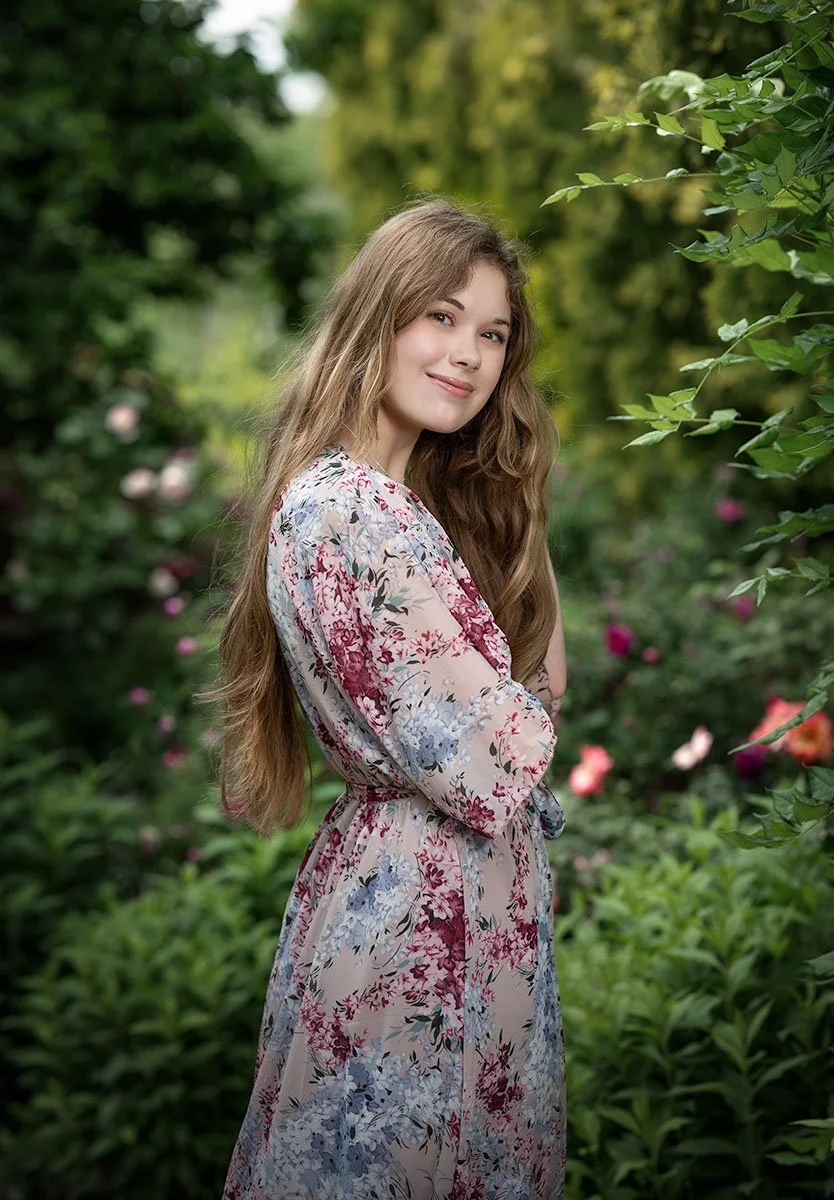 A young woman with long, wavy brown hair standing in a lush green garden with pink and white flowers, wearing a floral dress and smiling.