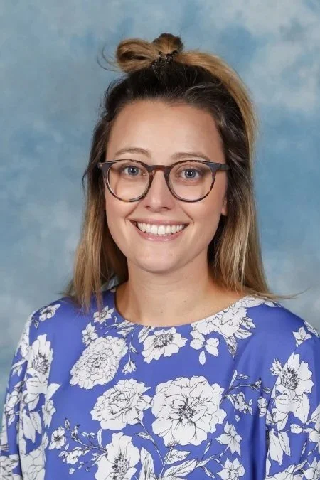 A young woman with glasses and a half-up hairstyle, wearing a blue floral top, smiling against a blue sky background.