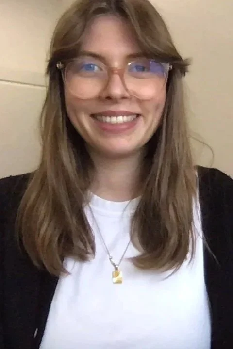 A young woman with long light brown hair and glasses, smiling, wearing a white shirt, black cardigan, and a gold necklace with a square pendant, standing against a beige wall.