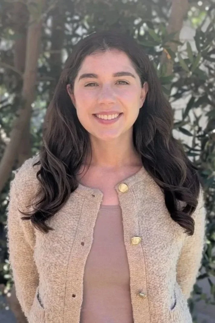 A young woman with long dark hair, smiling, wearing a beige textured jacket with gold buttons, standing outdoors in front of green foliage.