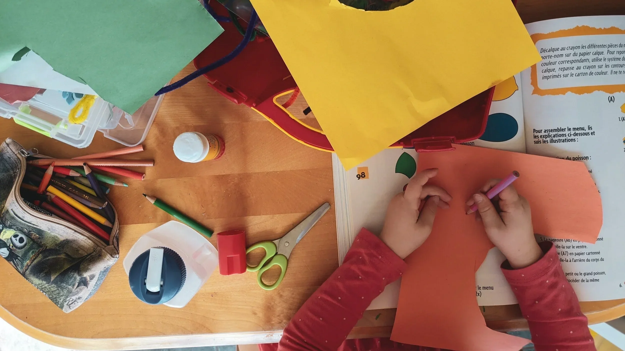 A child's hands coloring orange paper with a pink crayon at a wooden table, surrounded by school supplies including colored pencils, scissors, a glue stick, paper, and weeding tools.