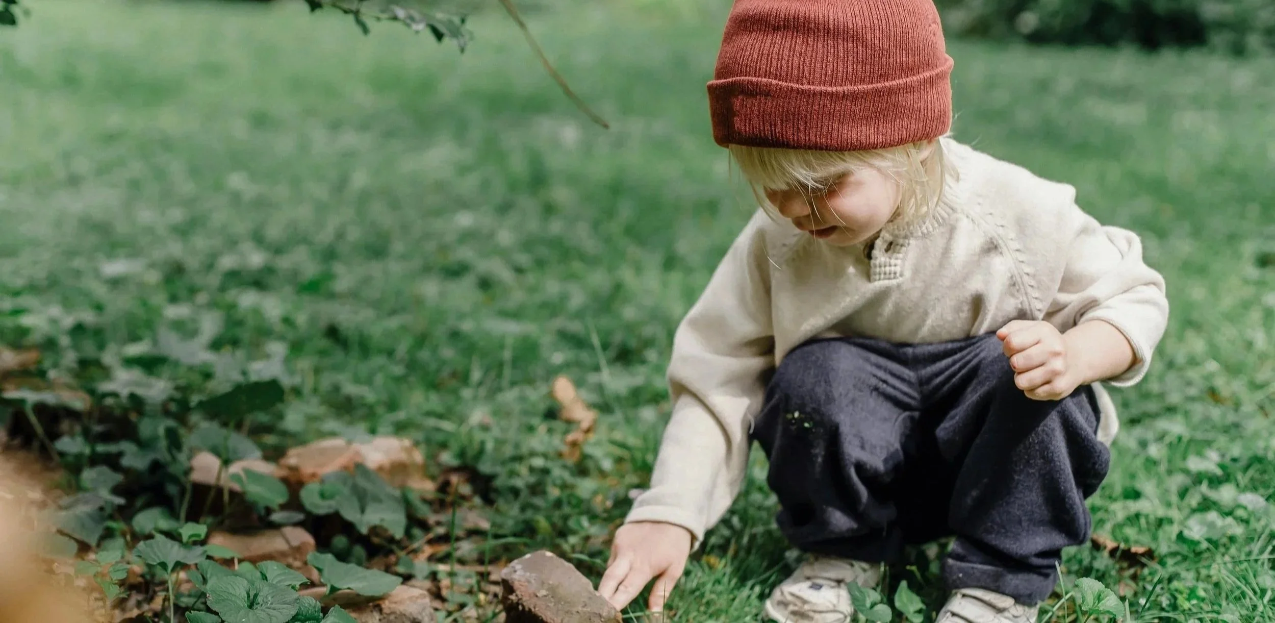 A young child wearing a red beanie, beige jacket, and dark pants crouches on grass, picking up a small rock or object near a patch of green plants.