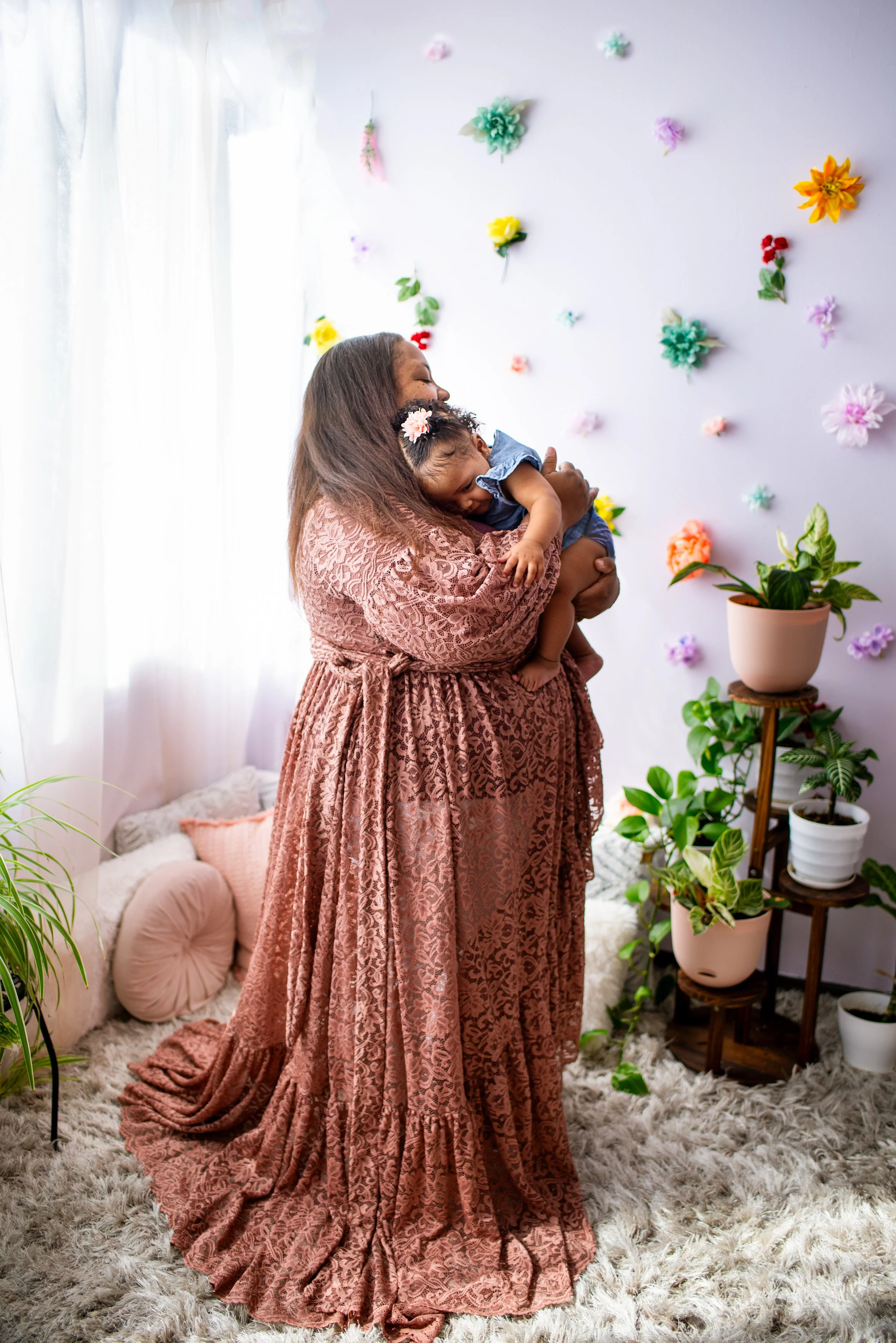 mom stands in pretty gown cuddling baby in front of flower wall