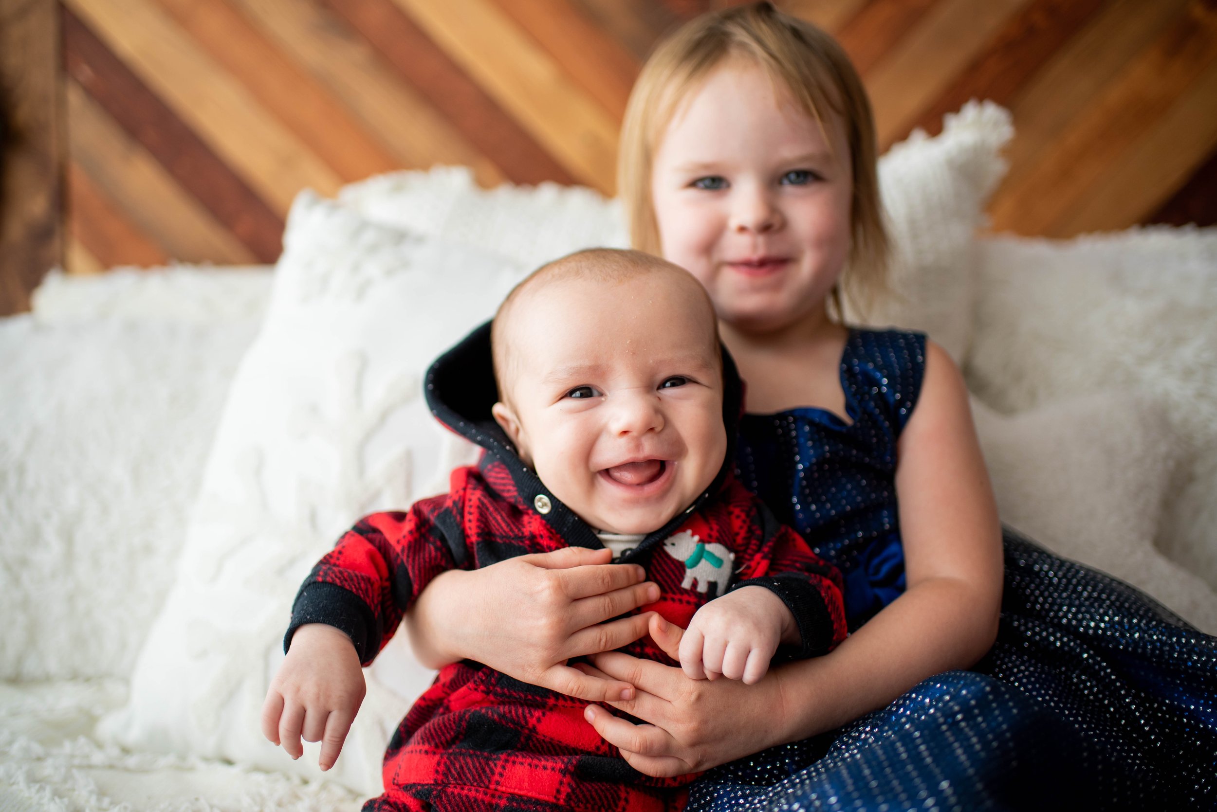 cute 2 month old grins wide for the camera while proud big sister toddler holds him for christmas