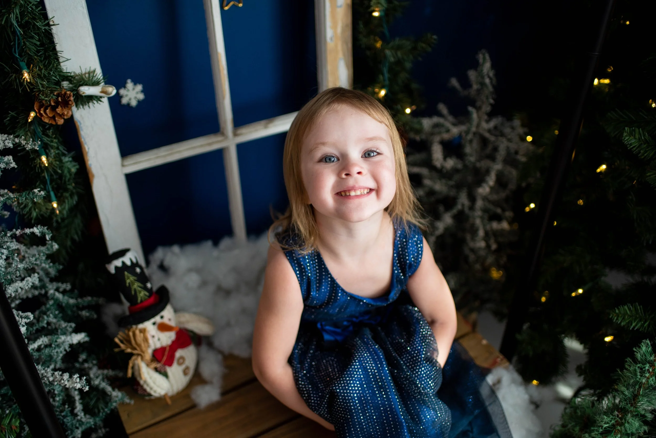3 year old girl sits in a blue dress smiling at the camera with christmas lights and cute snowmen