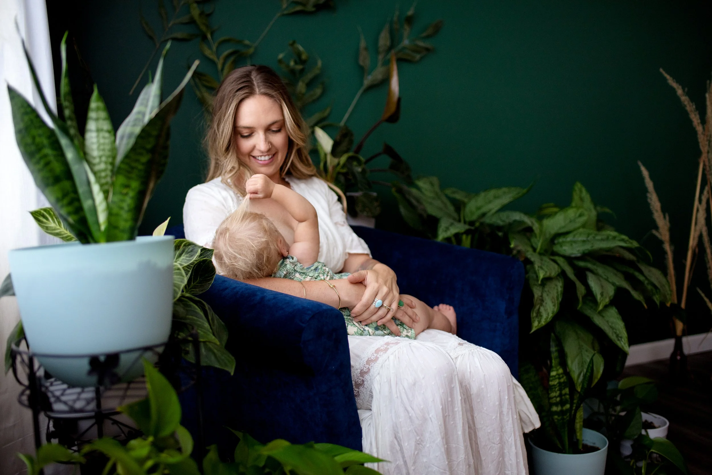 woman breastfeeding her toddler in a blue chair surrounded by plants indoors