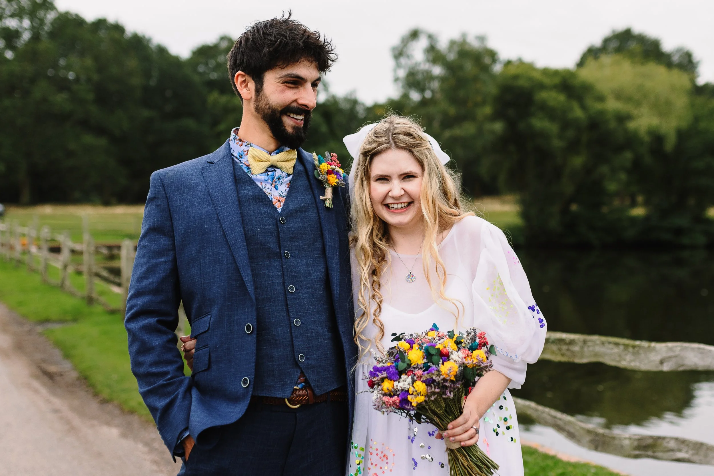 Bride and groom laughing together beside the lake at The York Club in Windsor, with the bride holding a colourful wildflower bouquet