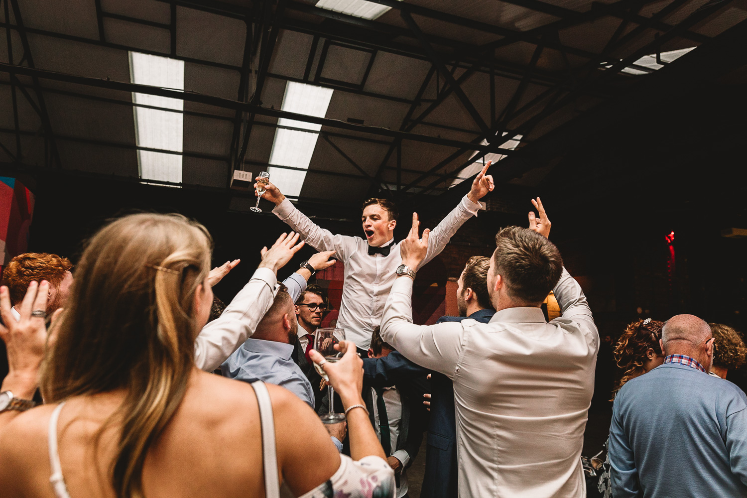 Photo shot from below of a groom on the shoulders of friends having fun with prosecco in one hand and arms in the air surrounded by friends in 92 Burton Road wedding venue in Sheffield