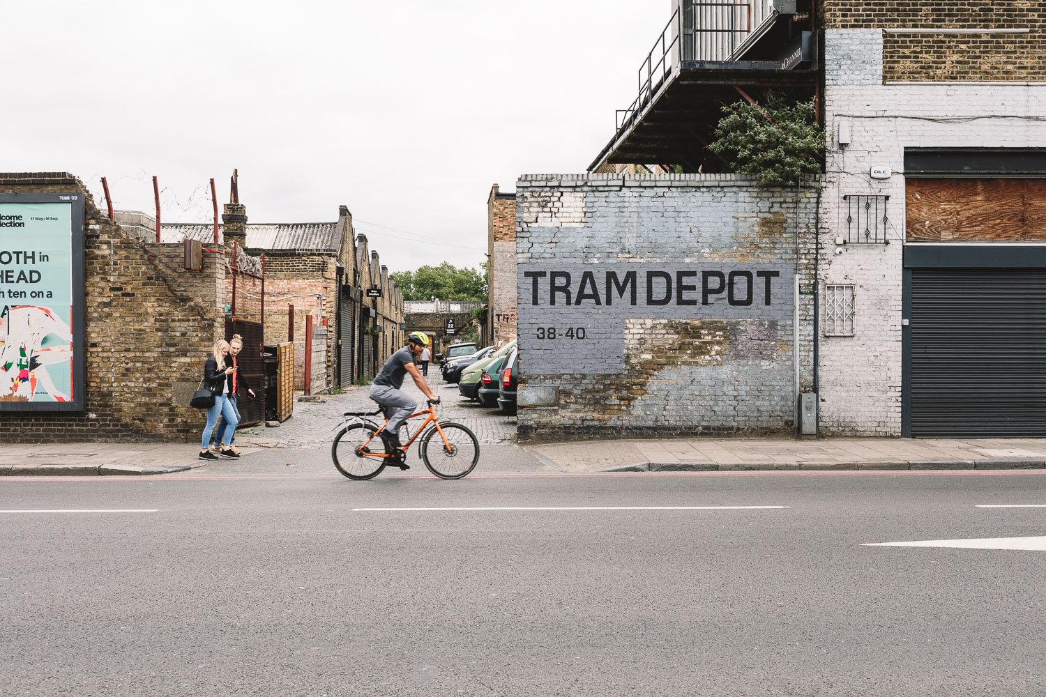 Exterior of Clapton Country Club Wedding Venue in London, brickwork with the words 'Tram Depot' painted on it leading to a line of warehouses, a cyclist and pedestrians go past the entrance.