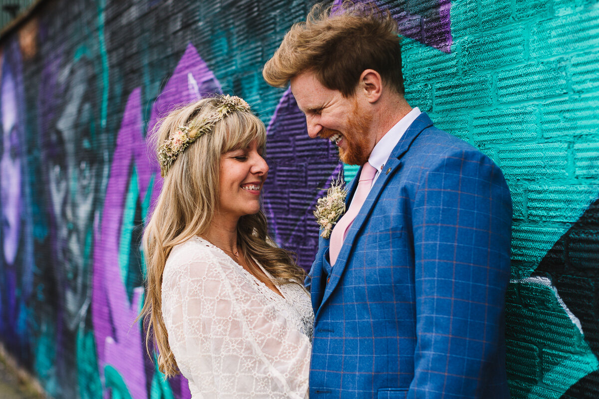 Boho bride with flower crown giggles with groom in blue suit with colourful graffti wall during relaxed couple portraits at Birmingham Wedding | Kate Jackson Photography-1.jpg