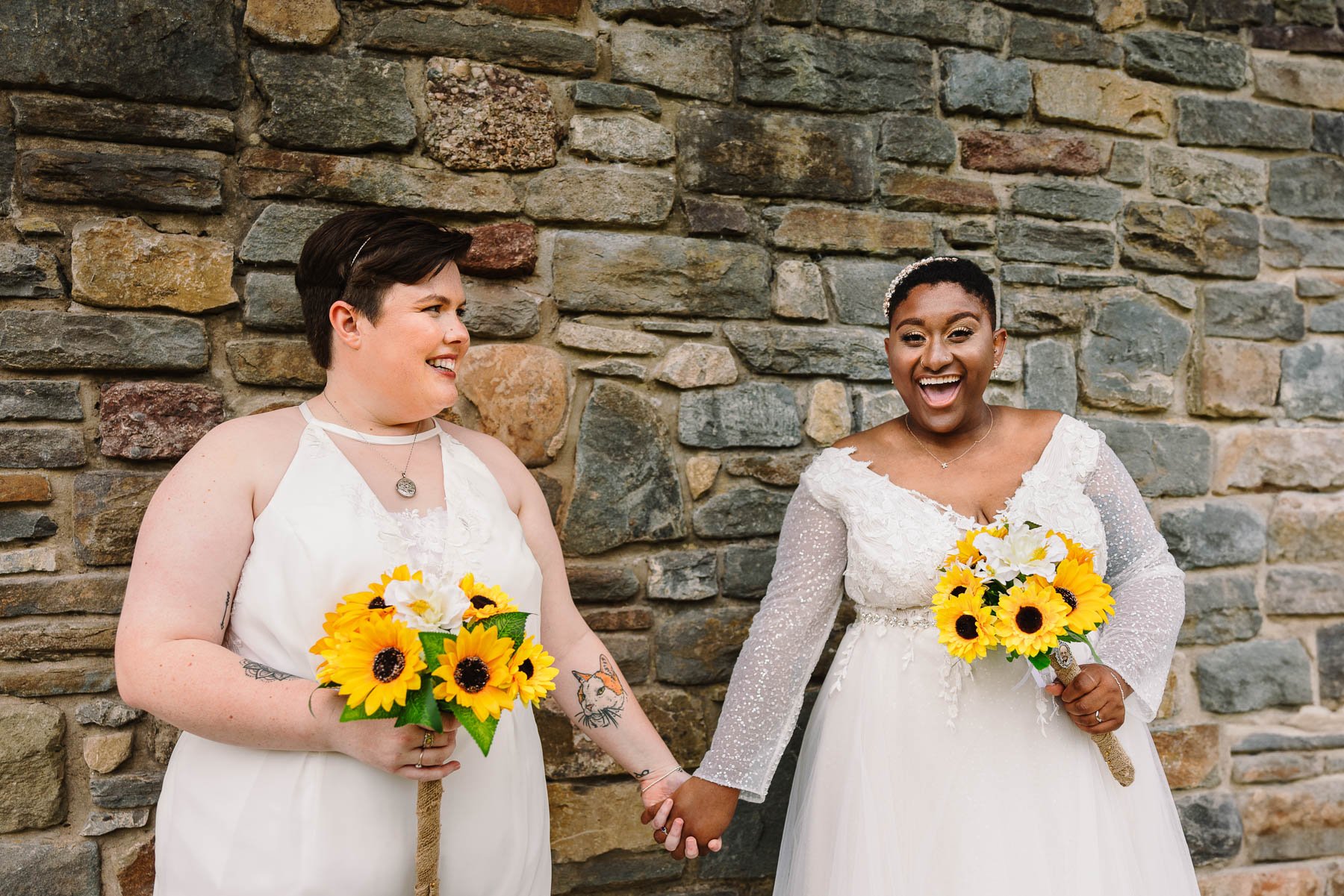 Two brides laughing and holding hands with sunflower bouquets at their summer wedding in Wales