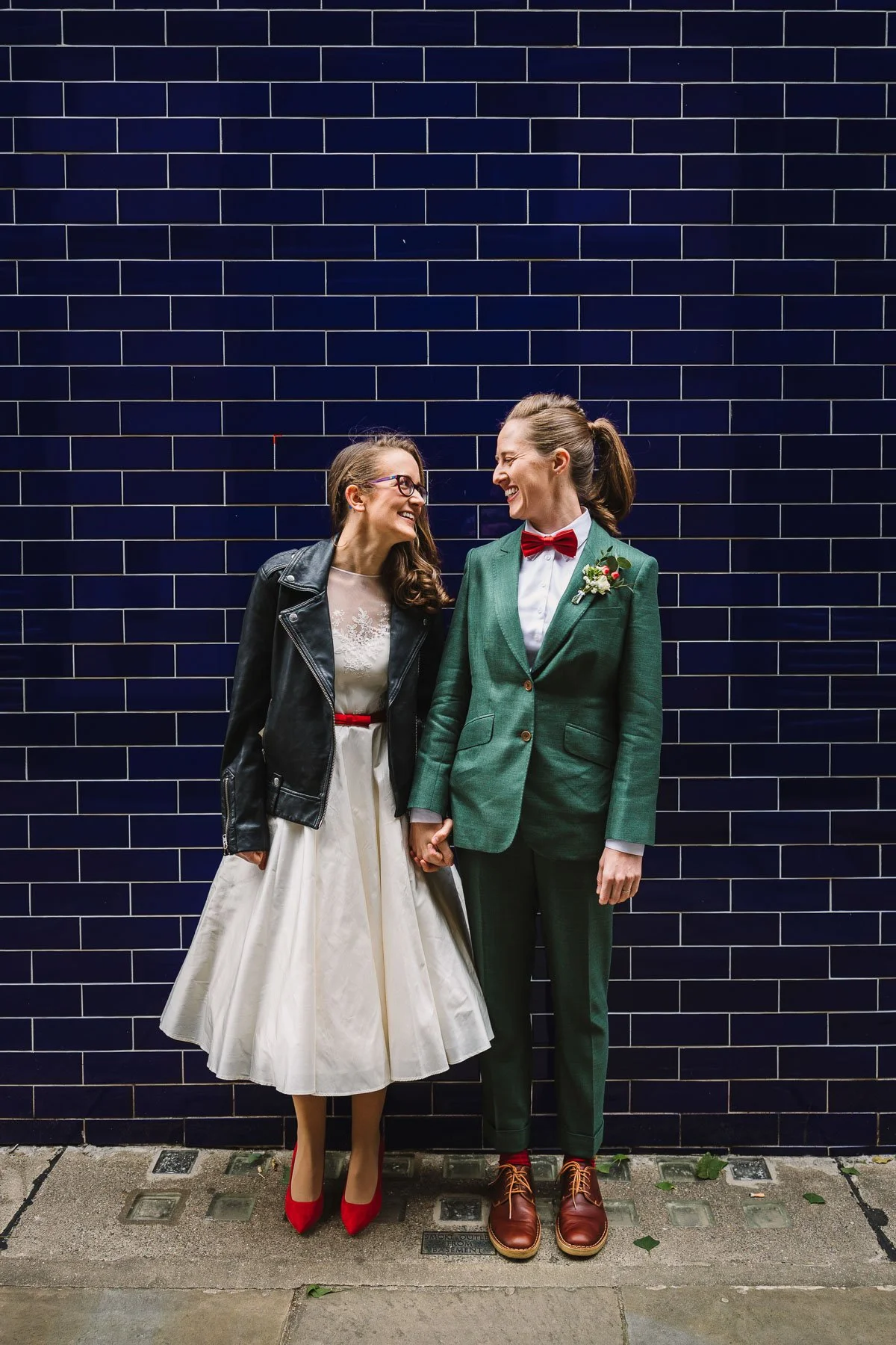 Two brides laughing and holding hands in front of a blue tiled wall during their London wedding