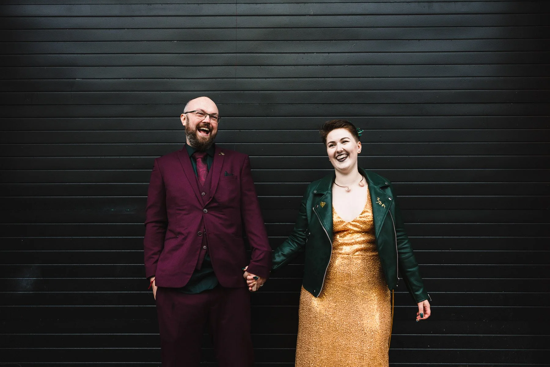 Laughing newlyweds holding hands in front of a black wall during relaxed wedding photos in Birmingham