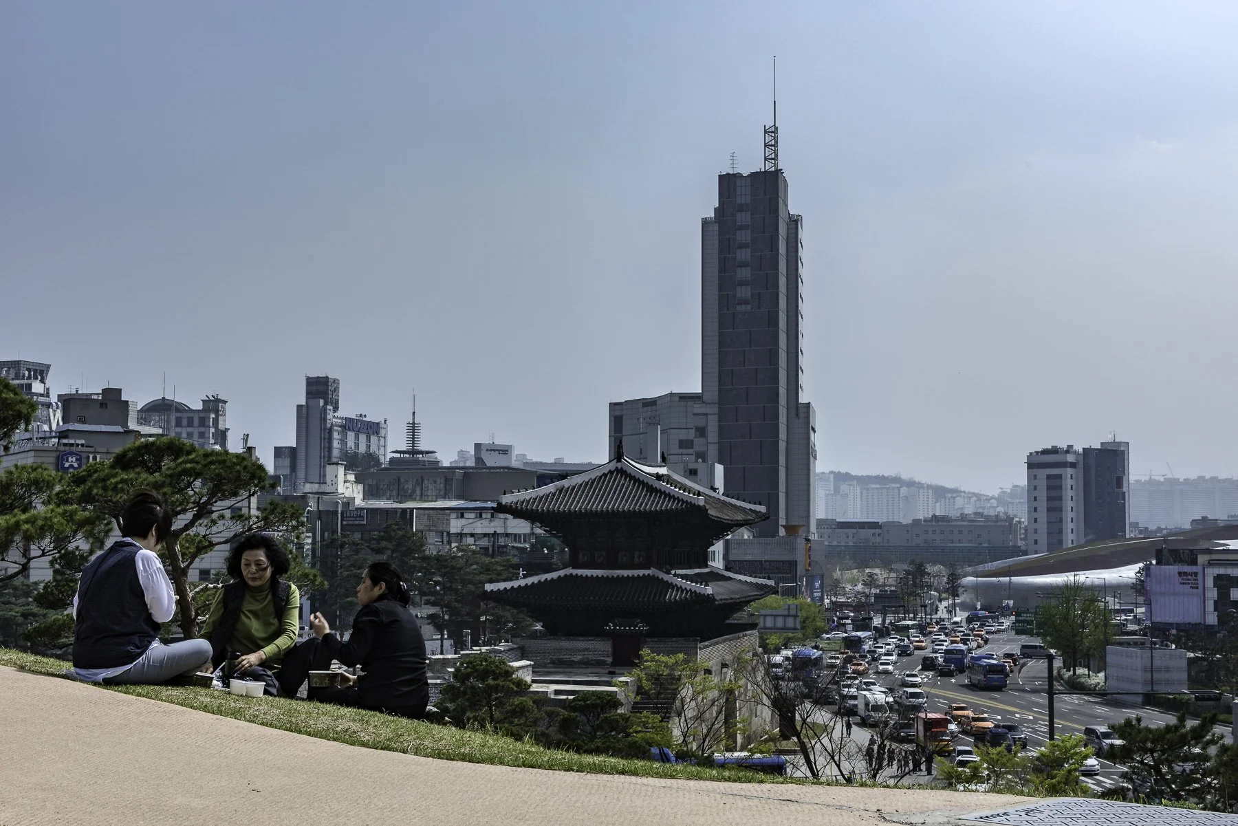 Three people sitting and talking on a park with traditional Korean architecture, high-rise buildings in the background, and a busy street below.
