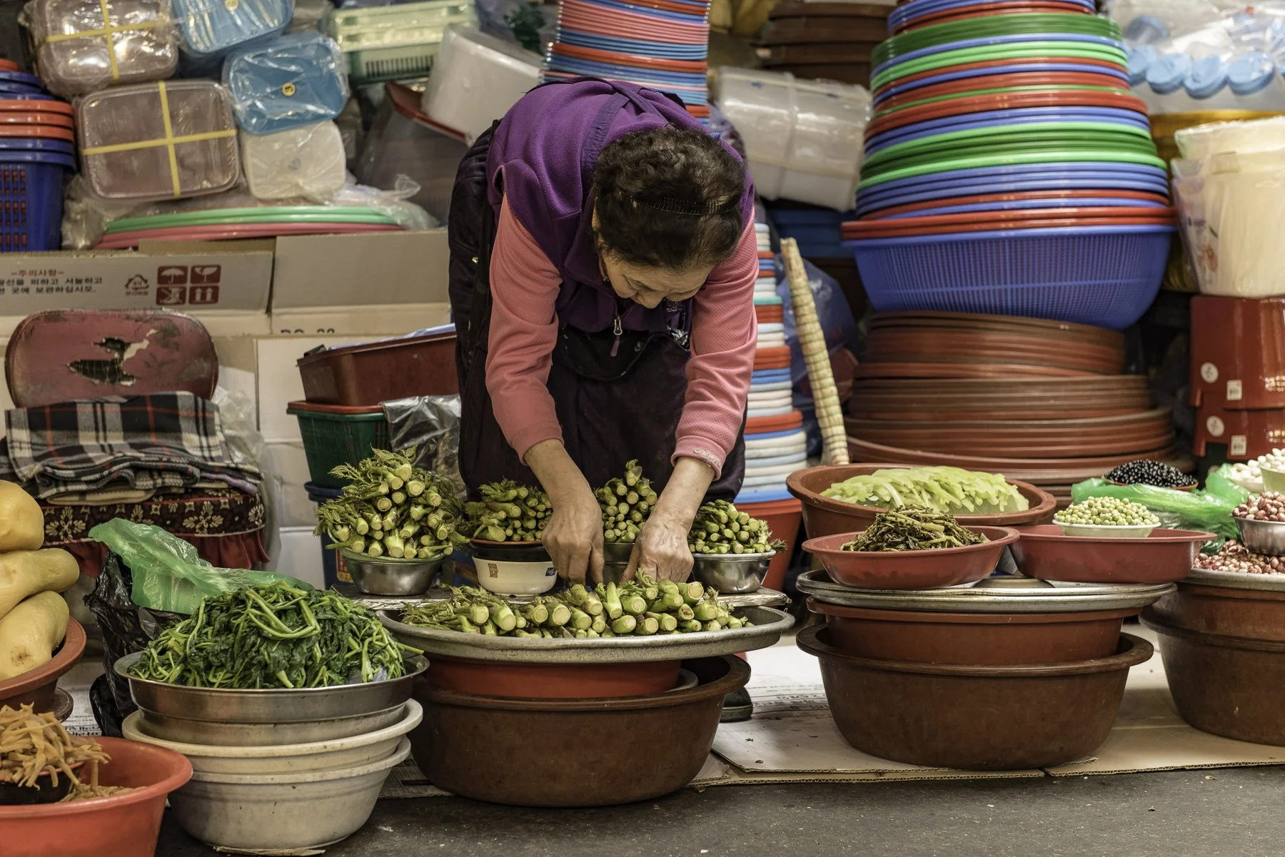 A woman arranging fresh vegetables at a market stall surrounded by colorful plastic bowls and containers.