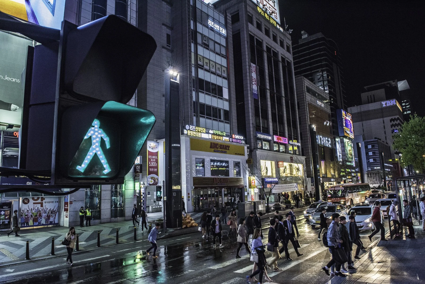 Nighttime city street with illuminated buildings, digital billboards, and a busy crosswalk. Pedestrians cross the wet street while cars and buses move along the road, and a green pedestrian light signals crossing.