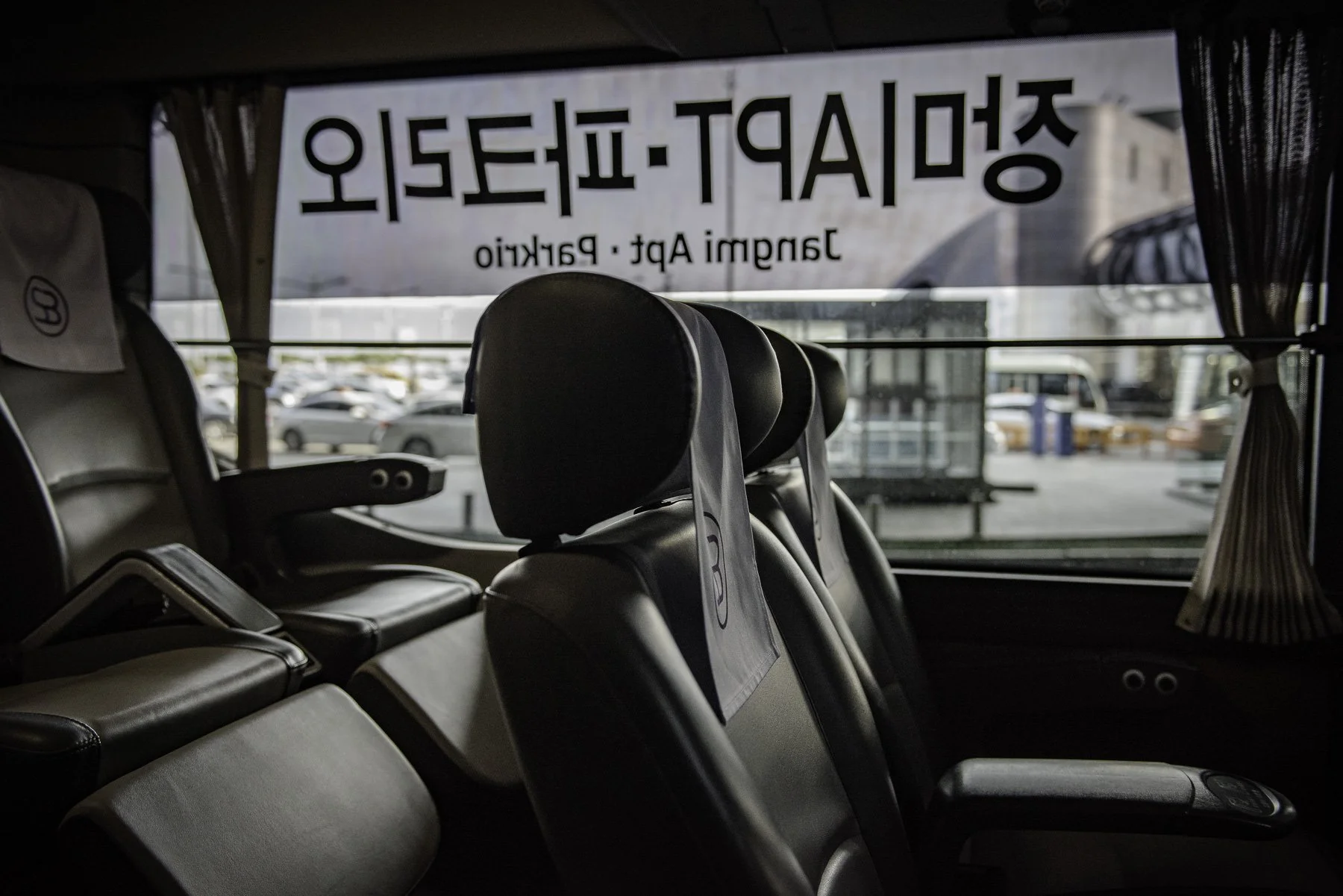 Interior of a bus showing empty black leather seats with headrests, with a window at the back displaying parked cars and a sign in Korean and English that reads 'SPORT-LEISAL' and 'Jangmi Apt. Parking'.