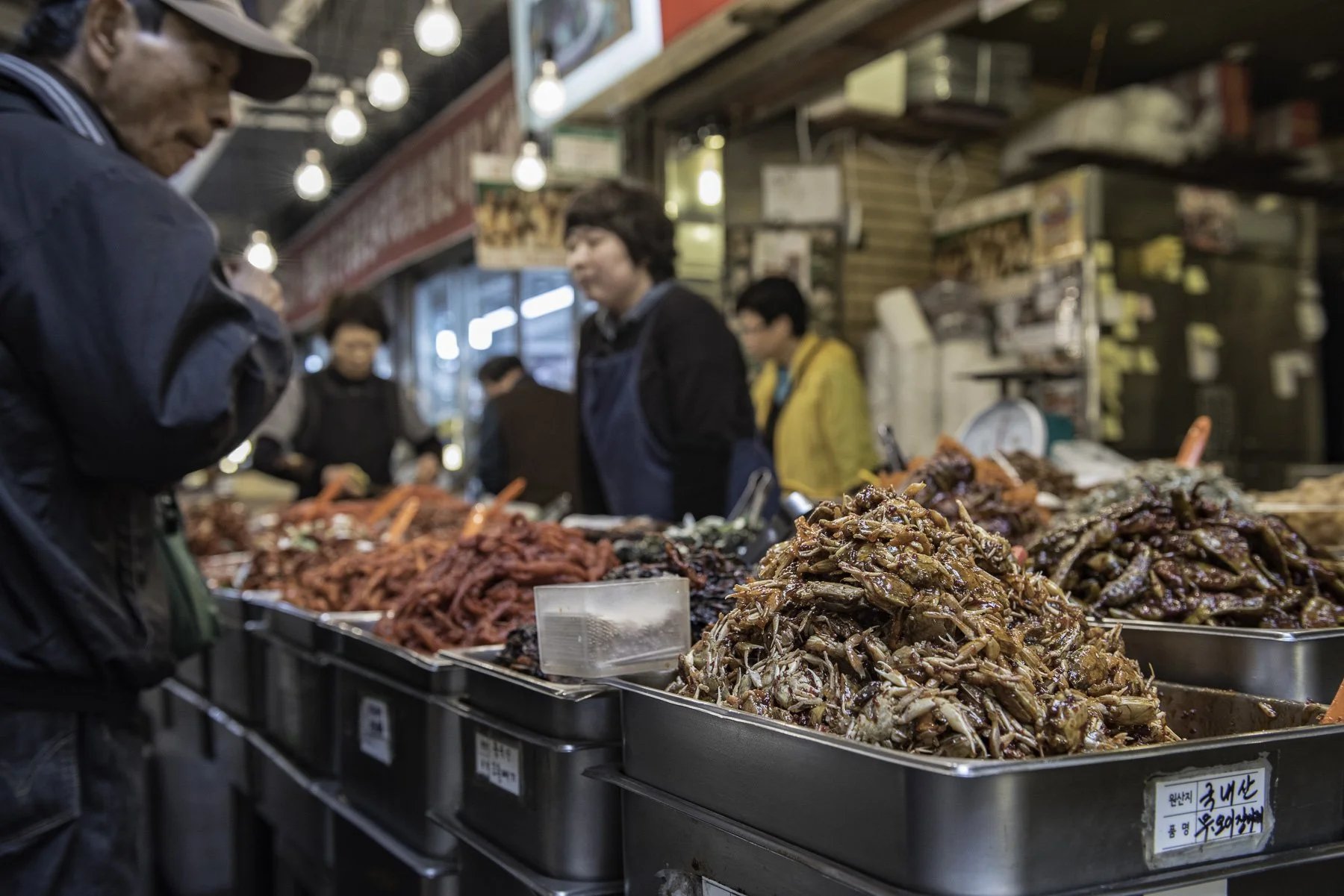 People shopping for various types of marinated and seasoned meat at a Korean market stall, with piles of seasoned meat in large metal trays in the foreground.